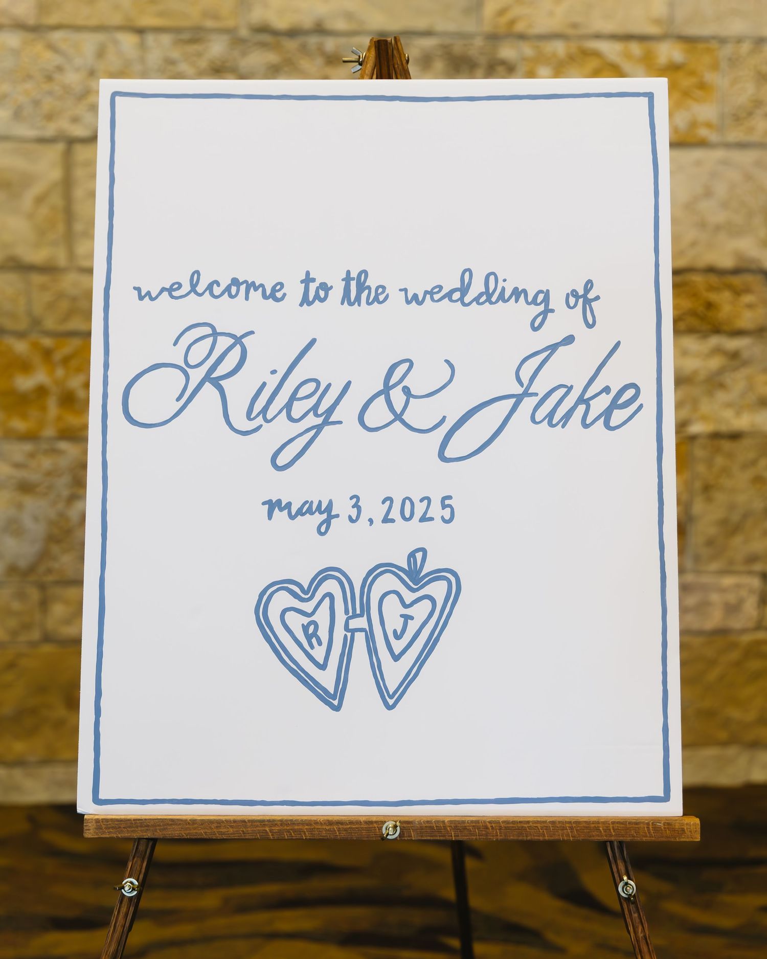 A wedding welcome sign featuring blue calligraphy for Riley & Jake's wedding on May 3, 2025 with two heart designs.
