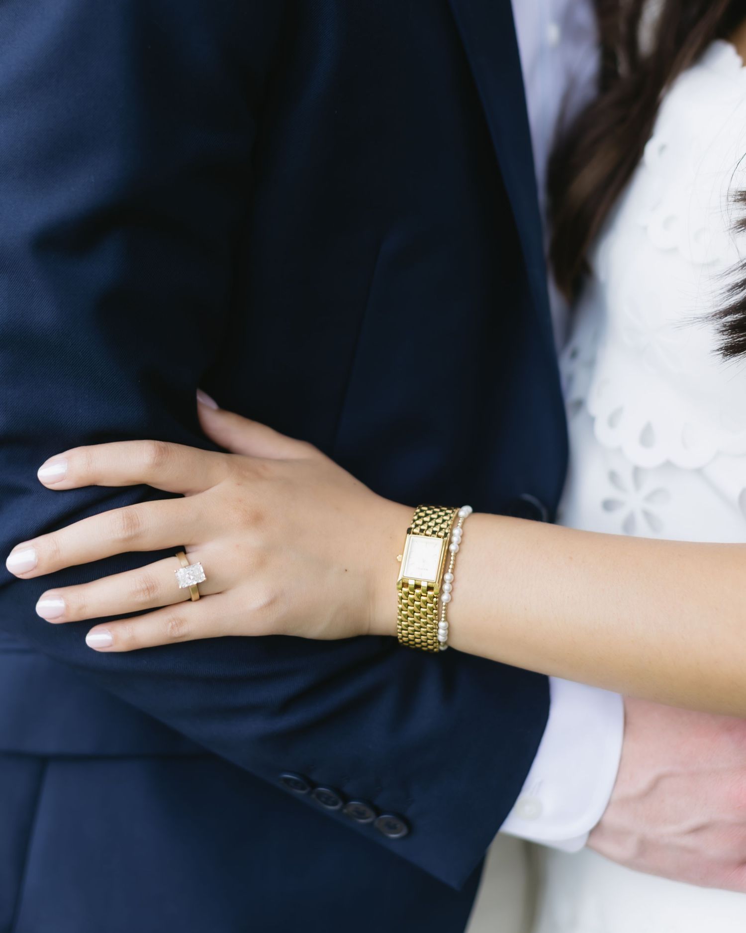 A hand wearing a gold watch and engagement ring reaches out to touch a dark suit jacket in a wedding photo.