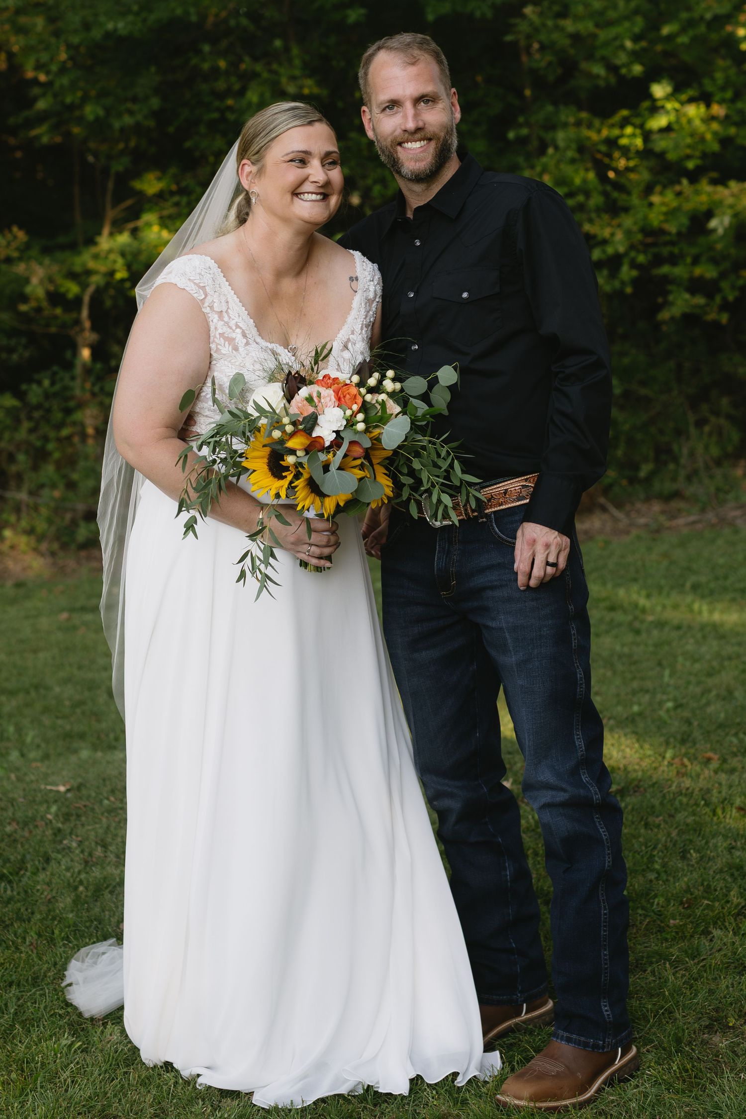 A couple poses outdoors on their wedding day with a sunflower and greenery bouquet.