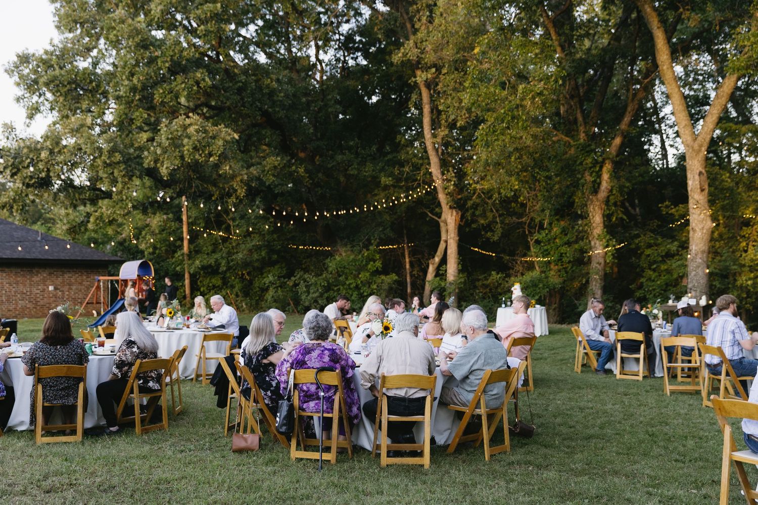 Panoramic view of outdoor wedding reception with wooden chairs arranged under mature trees.