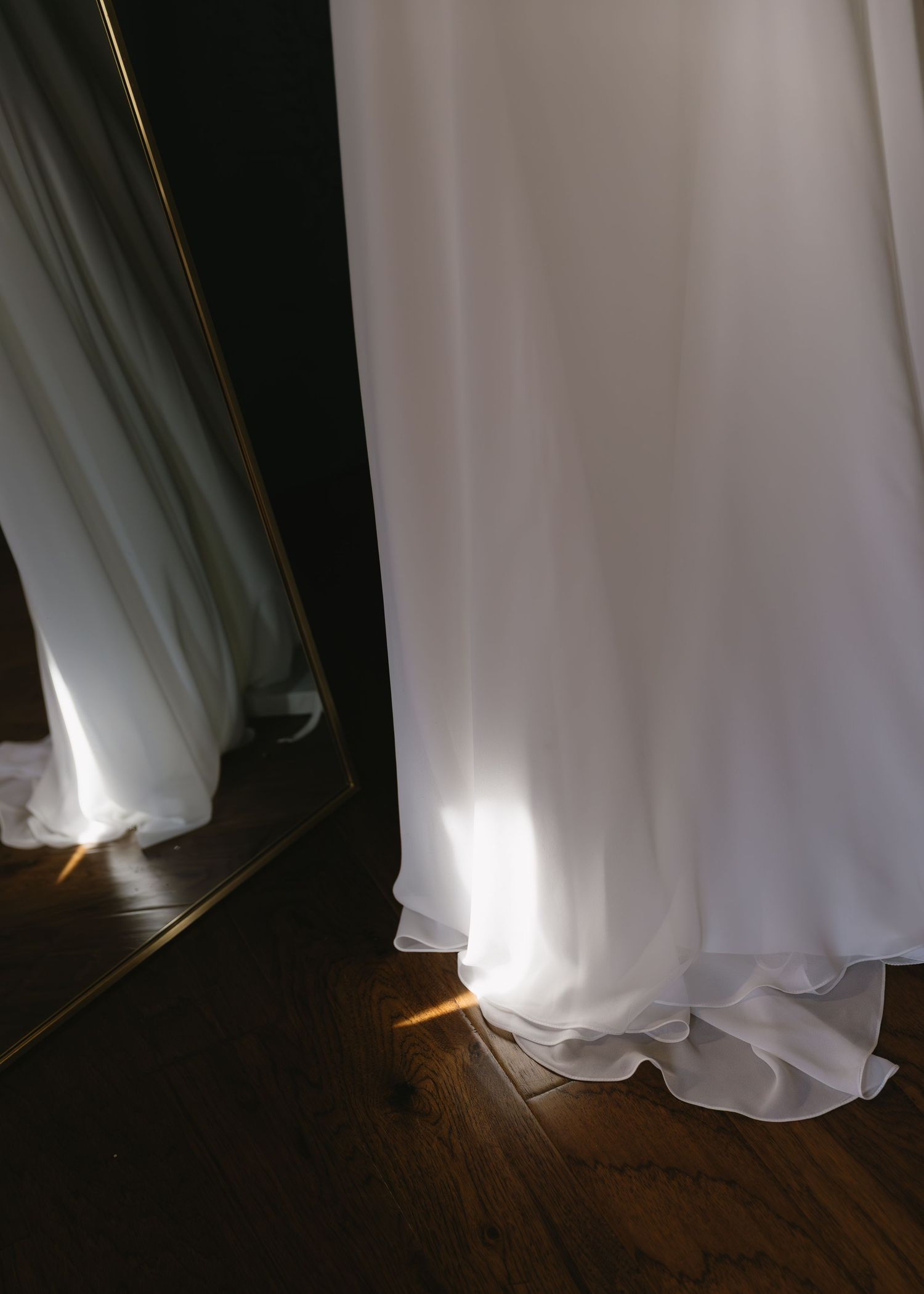 The bottom portion of a white wedding gown with flowing fabric shown against dark hardwood flooring.