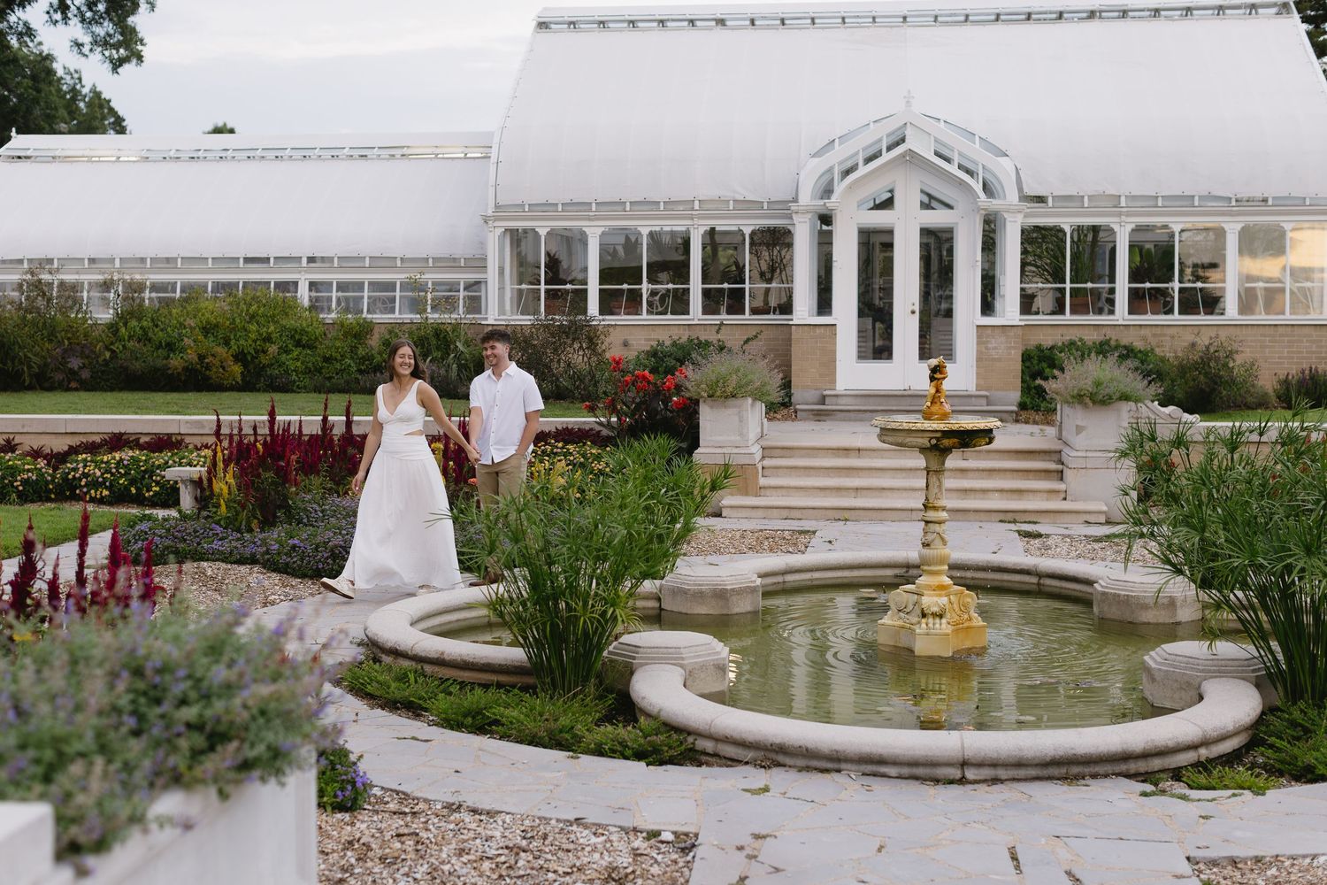 A peaceful garden setting with a decorative fountain and white pergola surrounded by manicured landscaping and pathways.