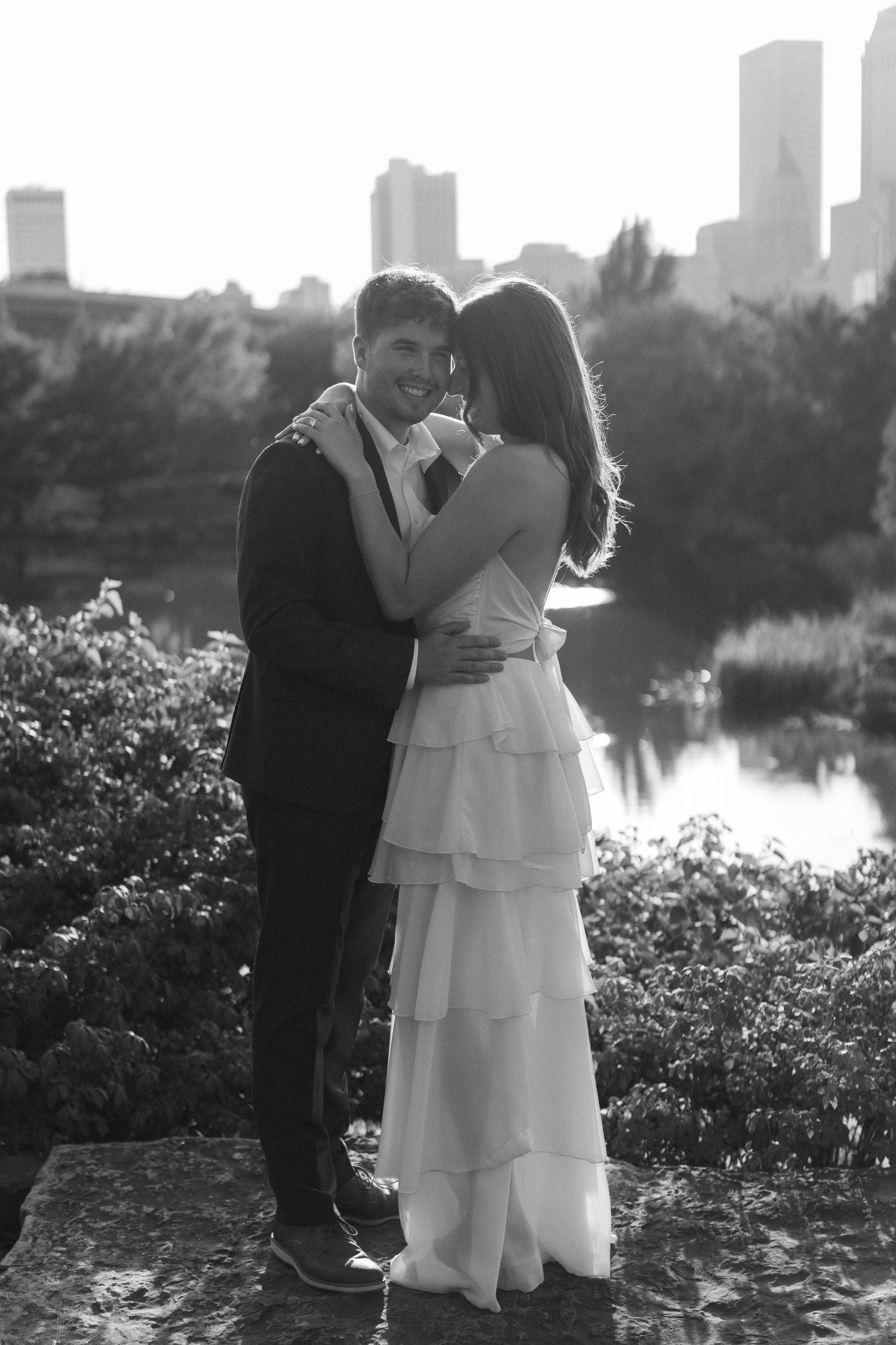 Wedding couple embracing near lakeside garden with city skyline in background during sunset.