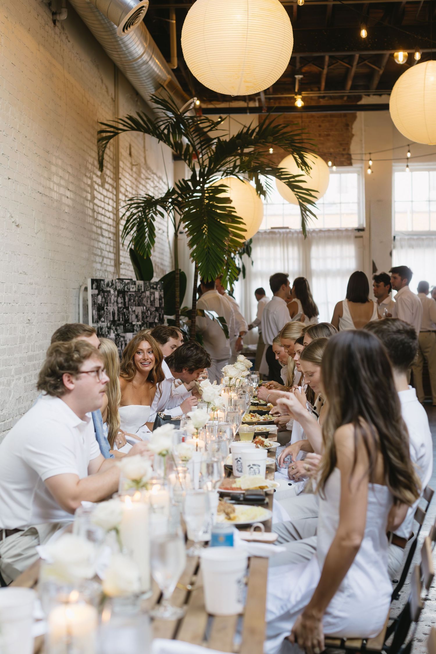 Reception dinner setup in white brick room with palm plants and round paper lanterns overhead.