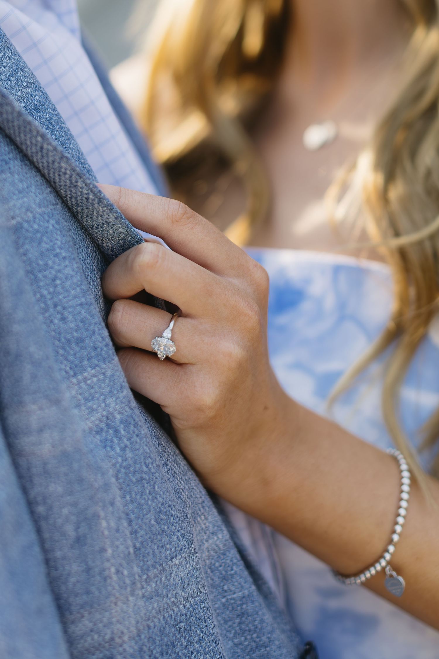 Close-up of an engagement ring and pearl bracelet with a blue blazer in soft lighting.