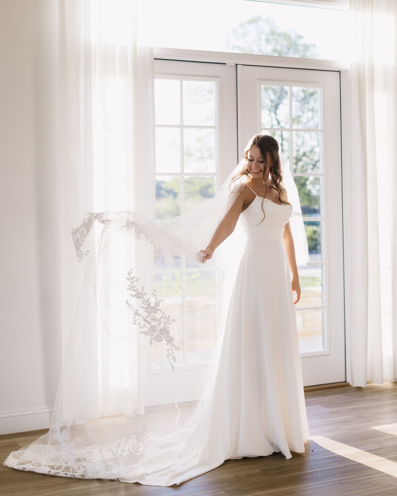 A bride in a flowing white dress stands by a sunlit window holding her long veil.