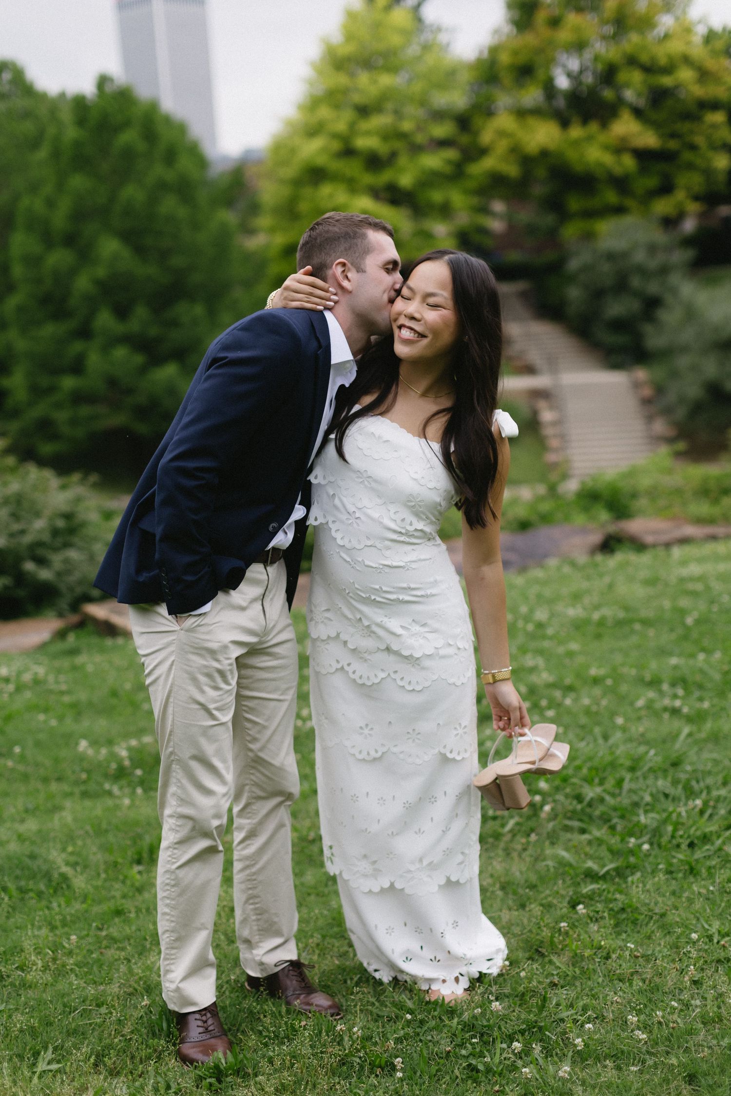 Couple shares a romantic moment on a grassy lawn surrounded by green trees and spring flowers.