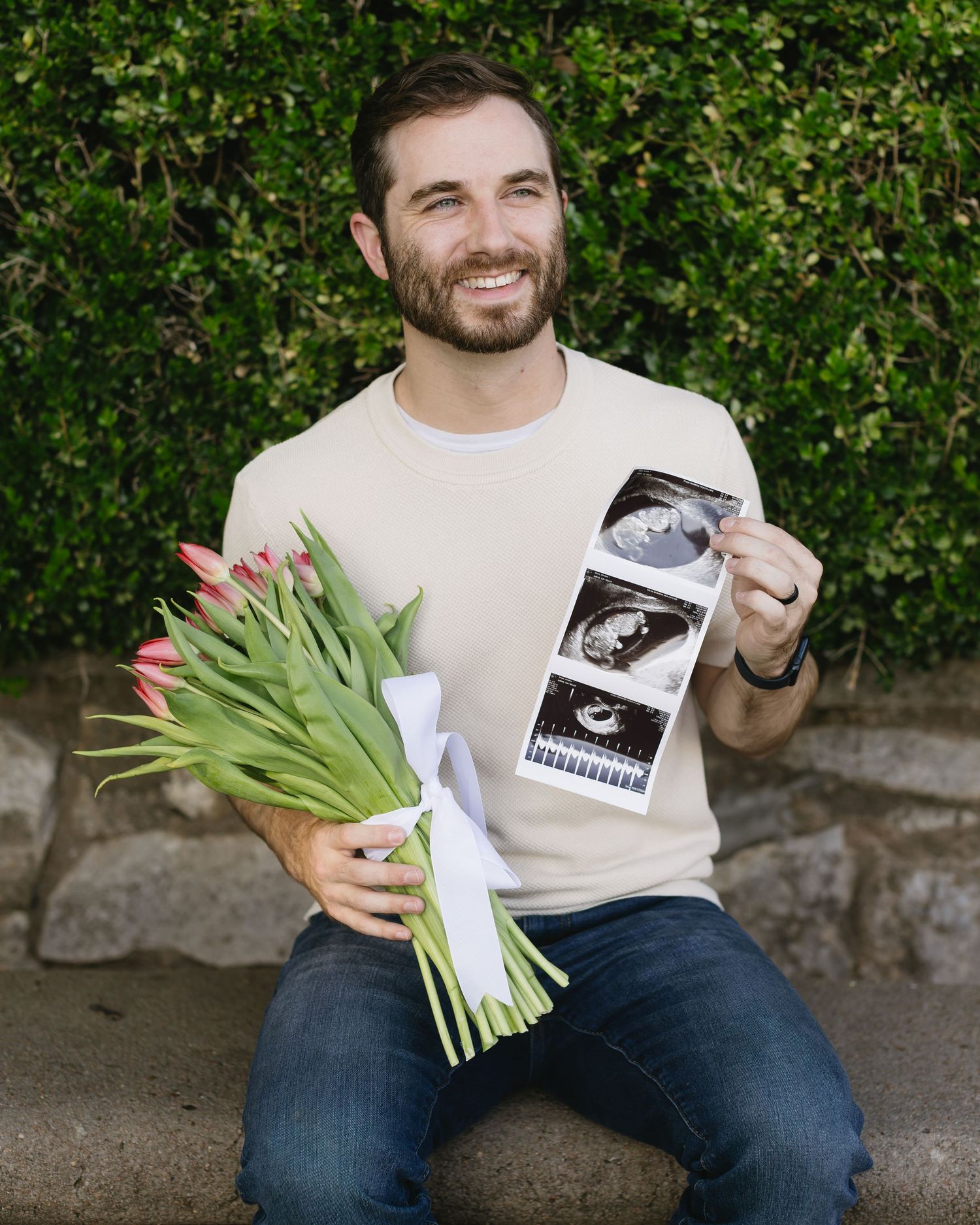 A person holding tulips and a sonogram photo sits on stone steps with leafy green backdrop.