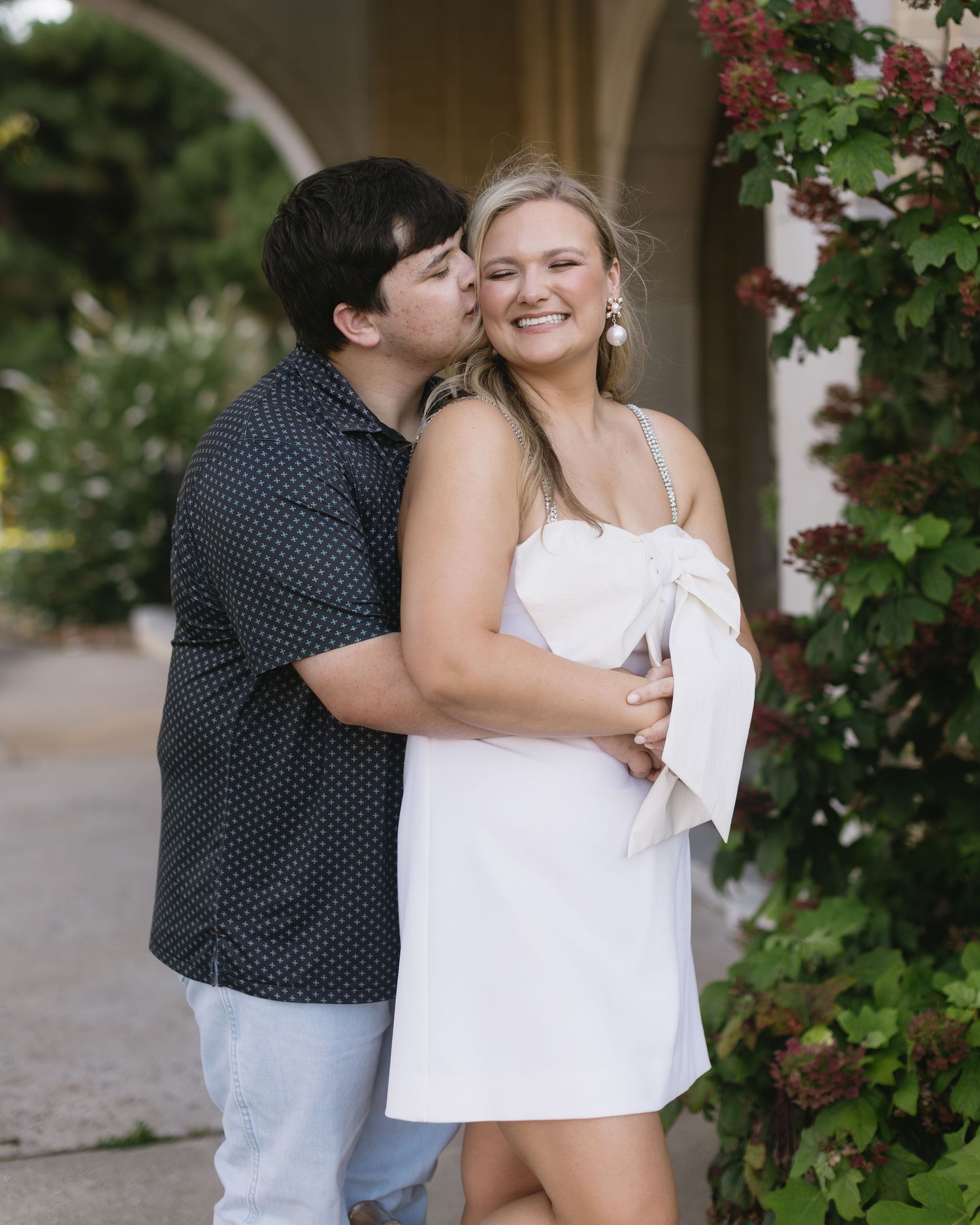 A romantic portrait showing affection between two people in front of blooming red flowers and greenery.