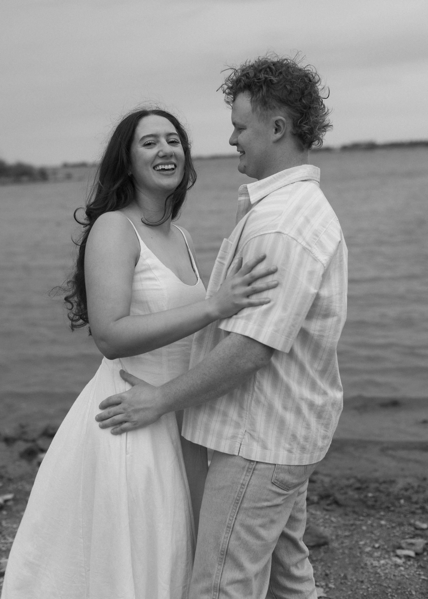 A couple in wedding attire embrace sweetly near the water in a serene romantic moment.