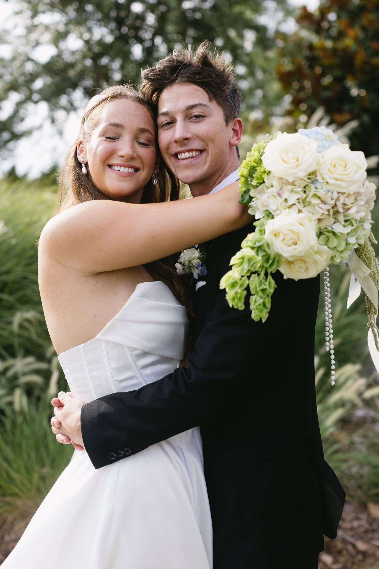 Romantic wedding couple embracing outdoors with white roses and greenery in their floral arrangement.