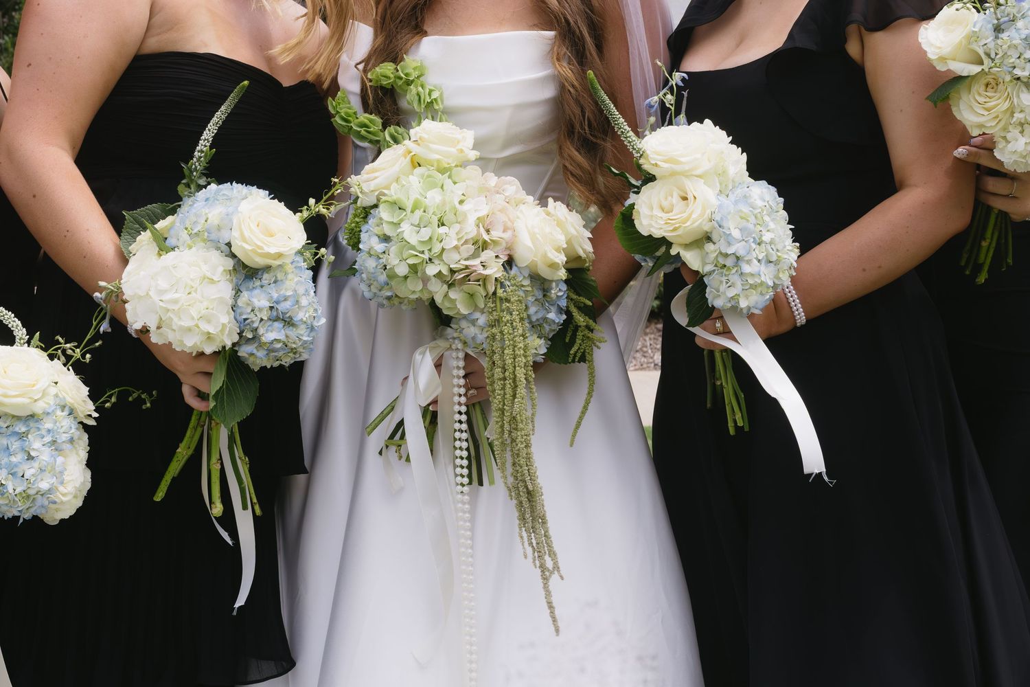 Row of white floral bouquets held by wedding party members in black dresses.