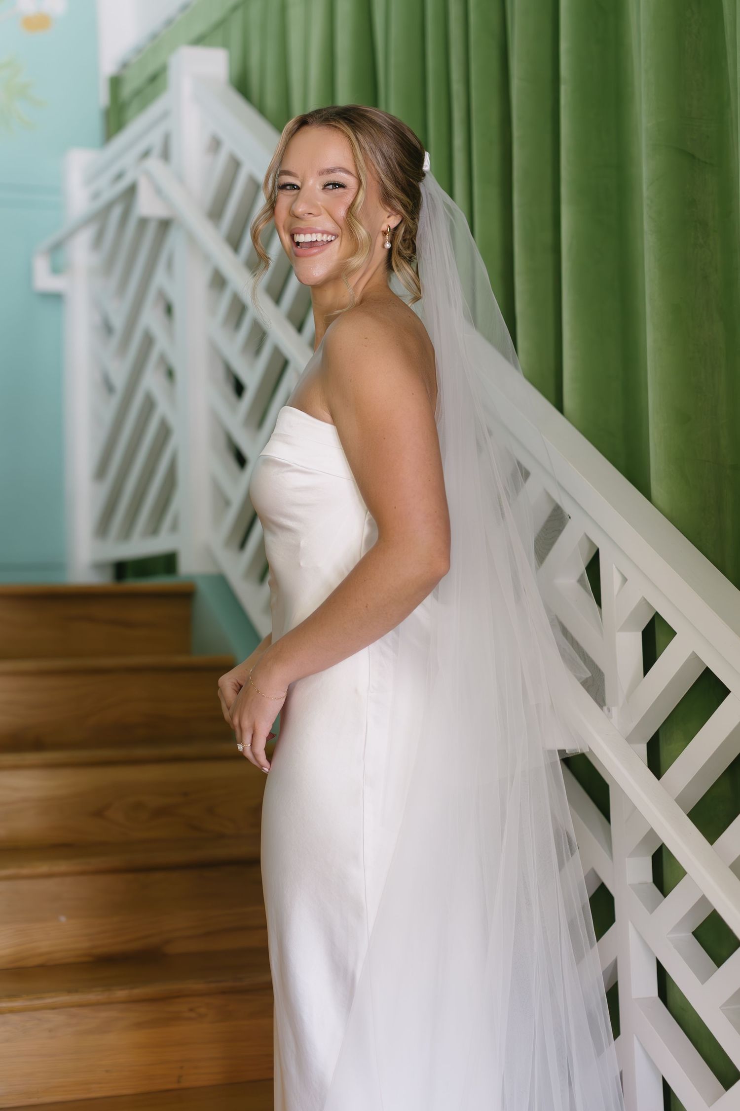 Bride in a sleeveless white wedding dress poses joyfully against a white lattice wall with green backdrop.