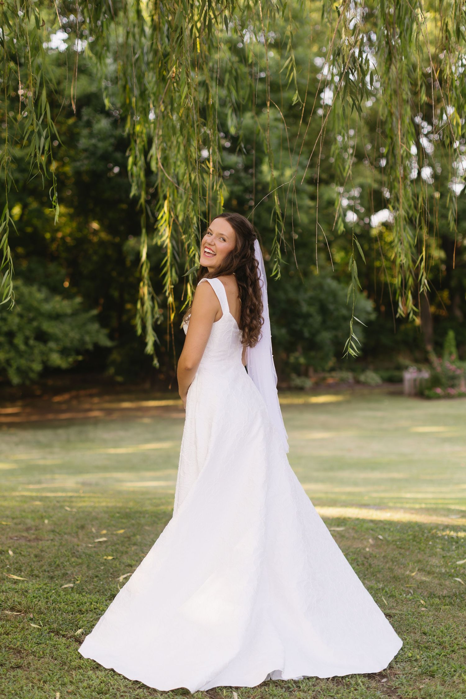 White wedding gown flowing elegantly under willow trees in a garden setting during golden hour.