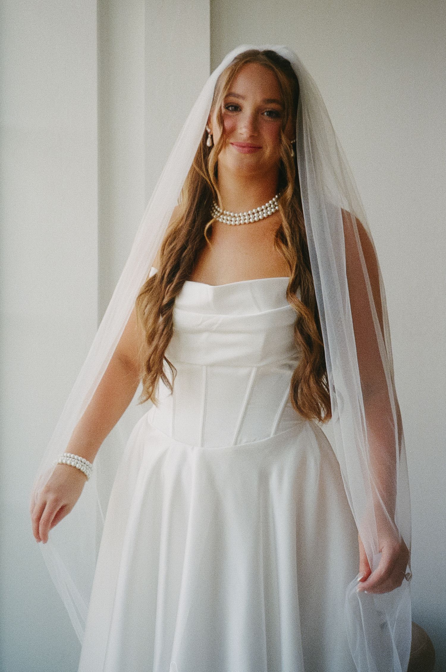 A bride in an elegant white strapless wedding gown with a flowing veil and pearl necklace stands by a window.
