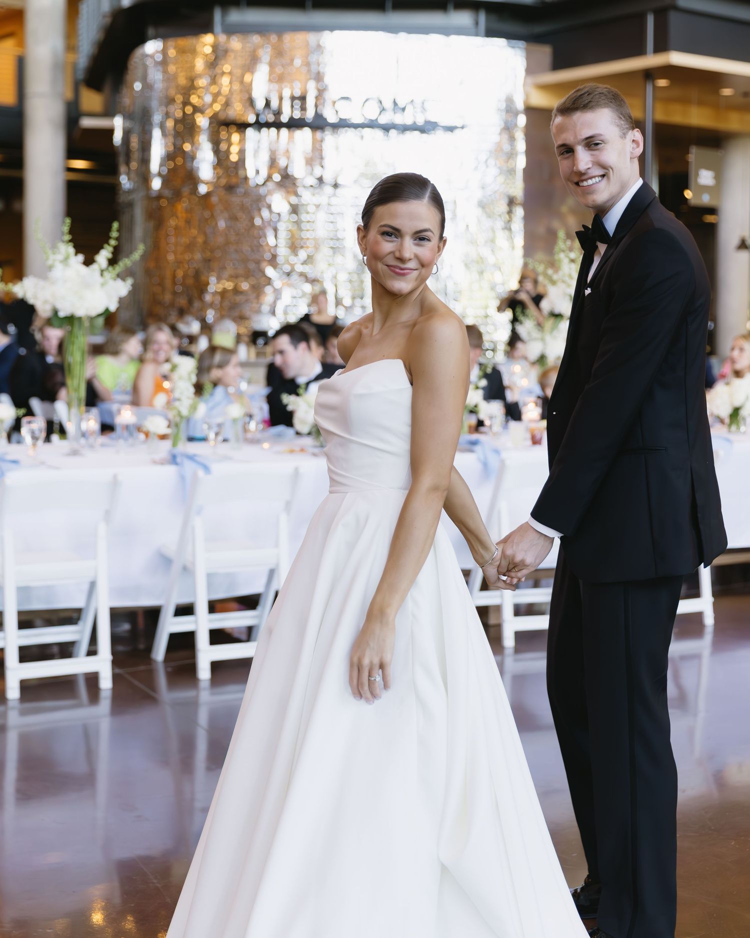 A reception venue decorated with white tables and chairs as newlyweds pose together.