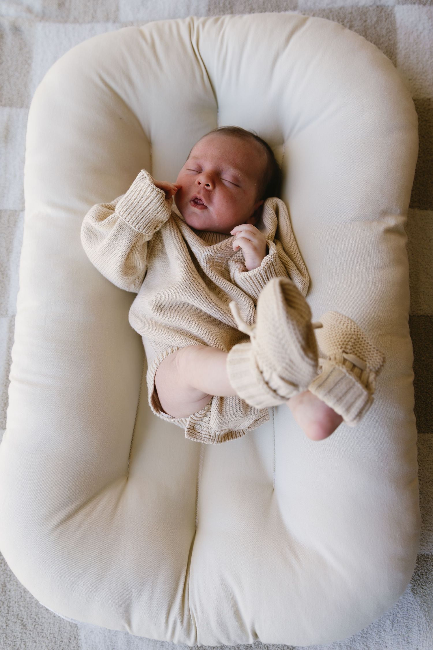 Newborn sleeping peacefully on white nursing pillow wearing cream knitted sweater.