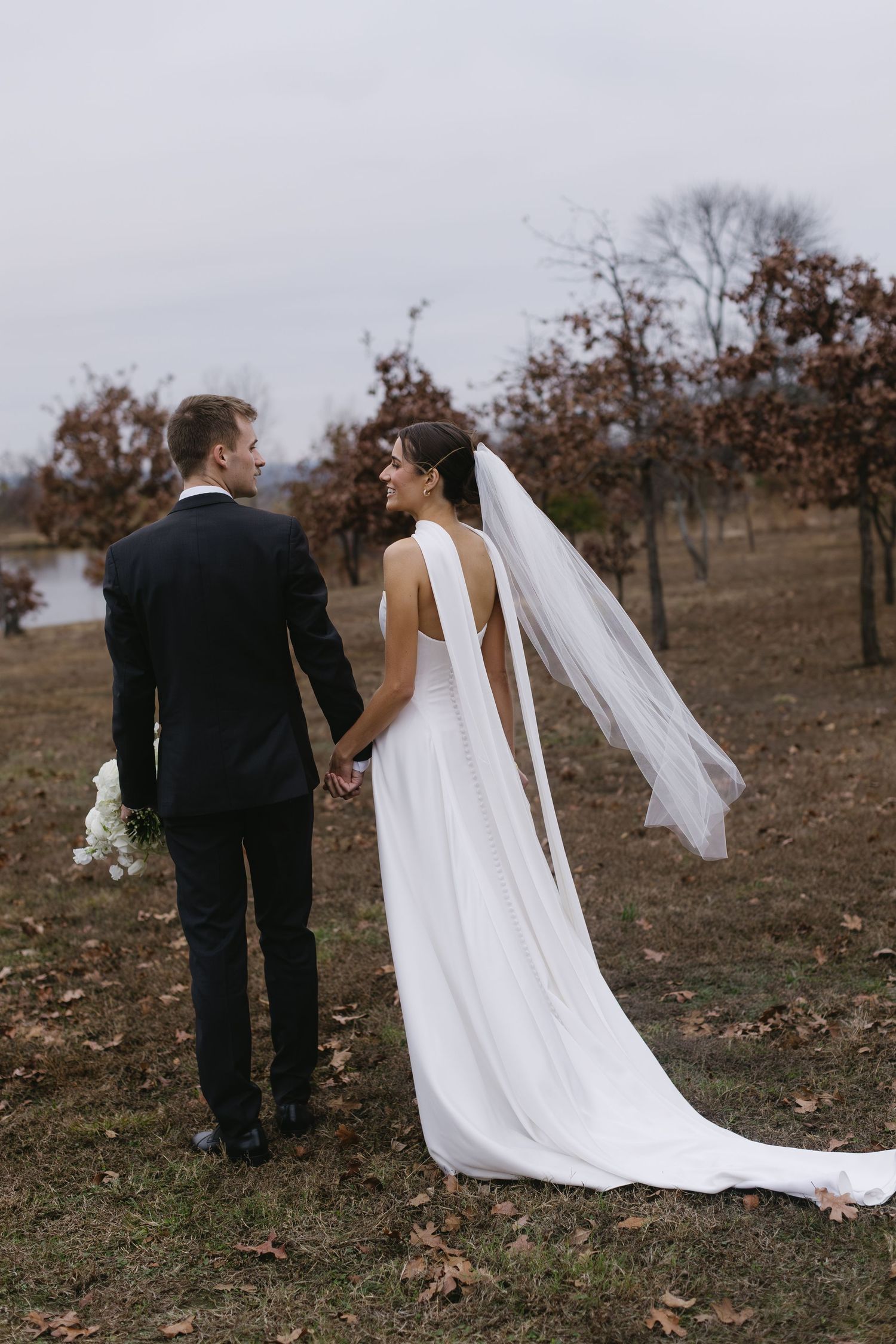 Newlyweds walk together across autumn field with bride's flowing white dress train trailing behind.