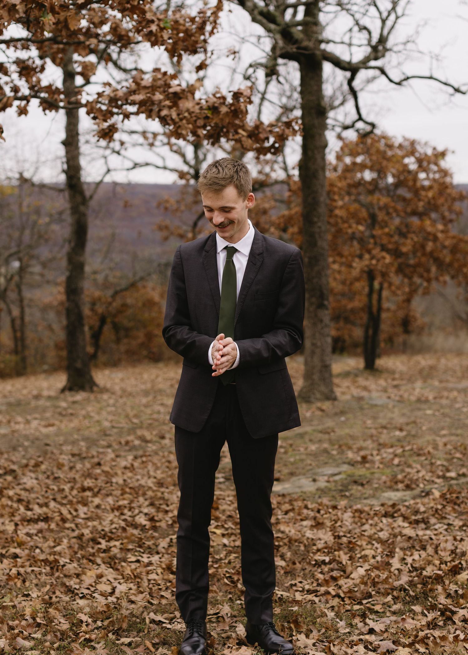 Person in dark suit standing on fallen autumn leaves in a wooded area.