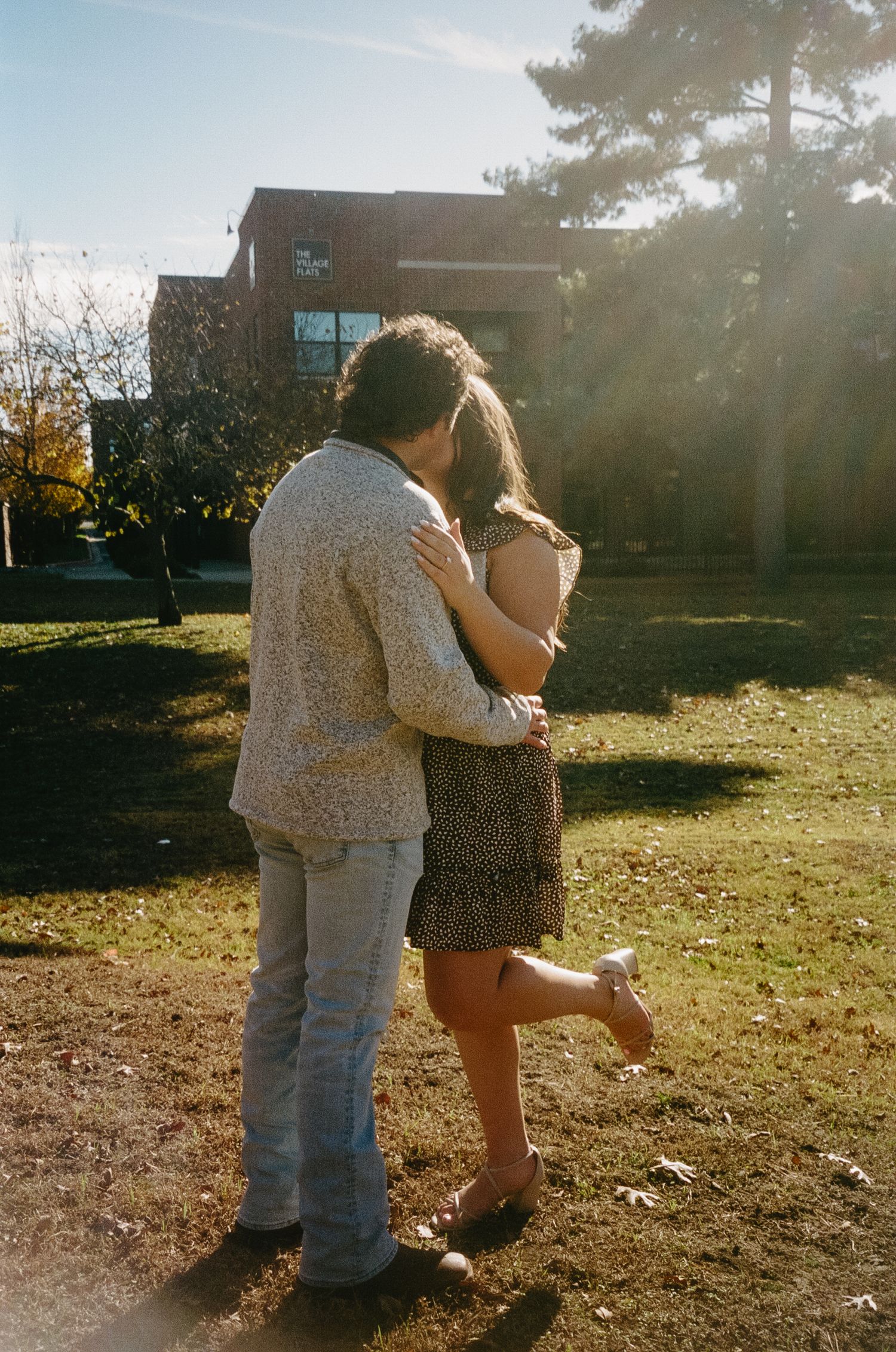 A couple embraces in a sunlit backyard on an autumn afternoon with a building visible in the background.