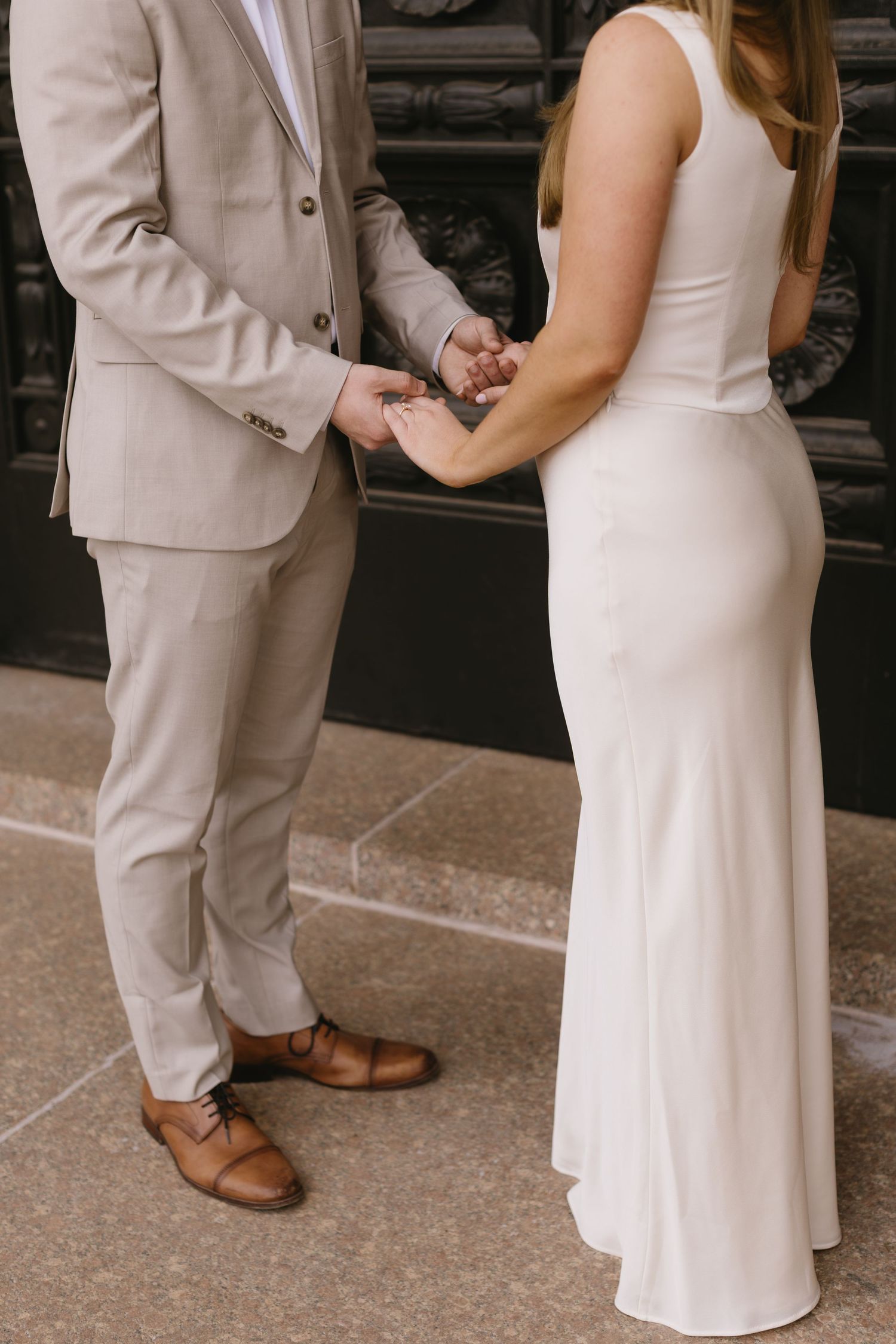 Close-up of couple holding hands, wearing beige suit and white dress with brown leather shoes.