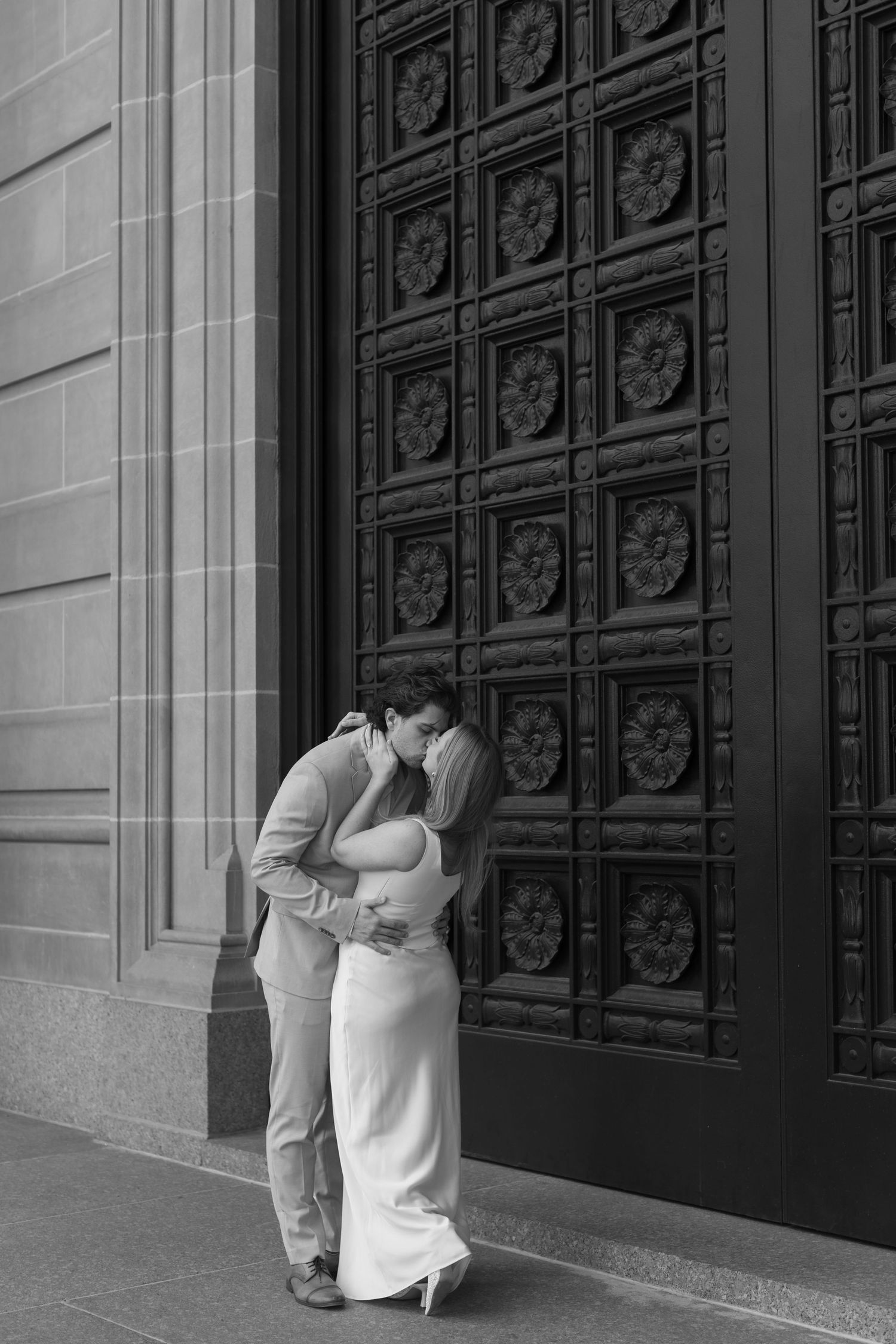 A couple shares an intimate moment in black and white against an ornate door at a historic building.