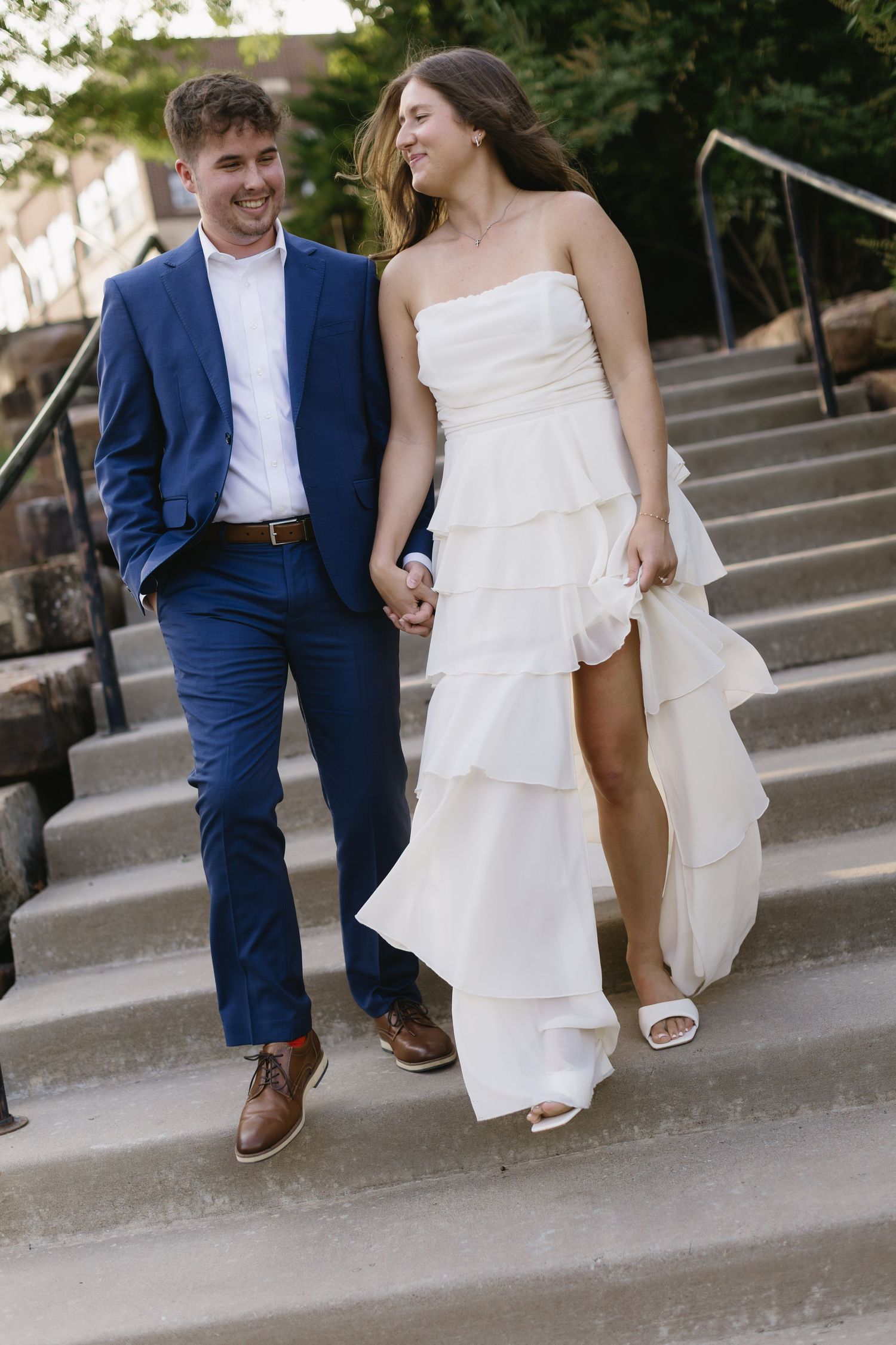 A couple walks down stone steps together, with the bride wearing a flowing white dress and groom in navy suit.