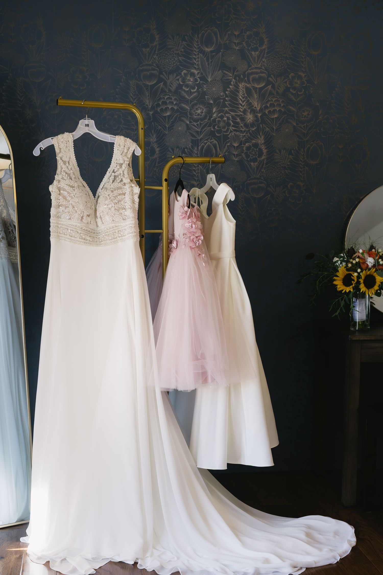 Wedding dresses and flower girl dresses hanging on display against a dark wall with delicate lace and tulle details.