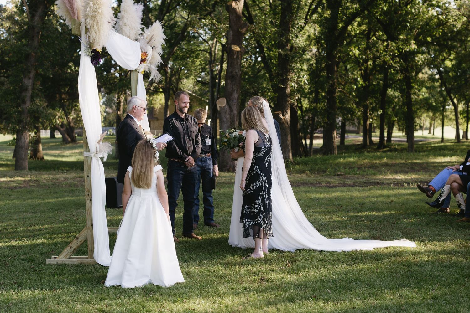 An outdoor wedding ceremony under a white draped arch surrounded by trees.