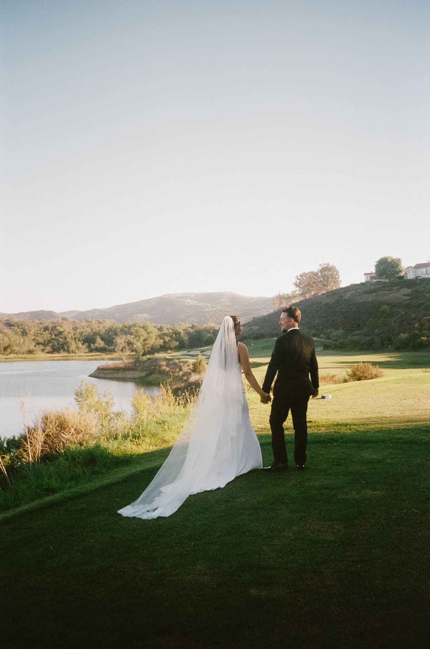 A scenic wedding photo overlooking a lake with rolling hills, featuring a couple in formal attire at sunset.