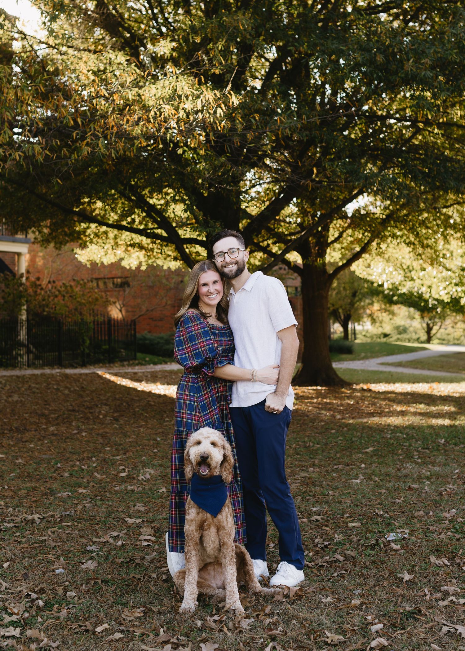 A couple poses with their goldendoodle dog under trees with autumn foliage.