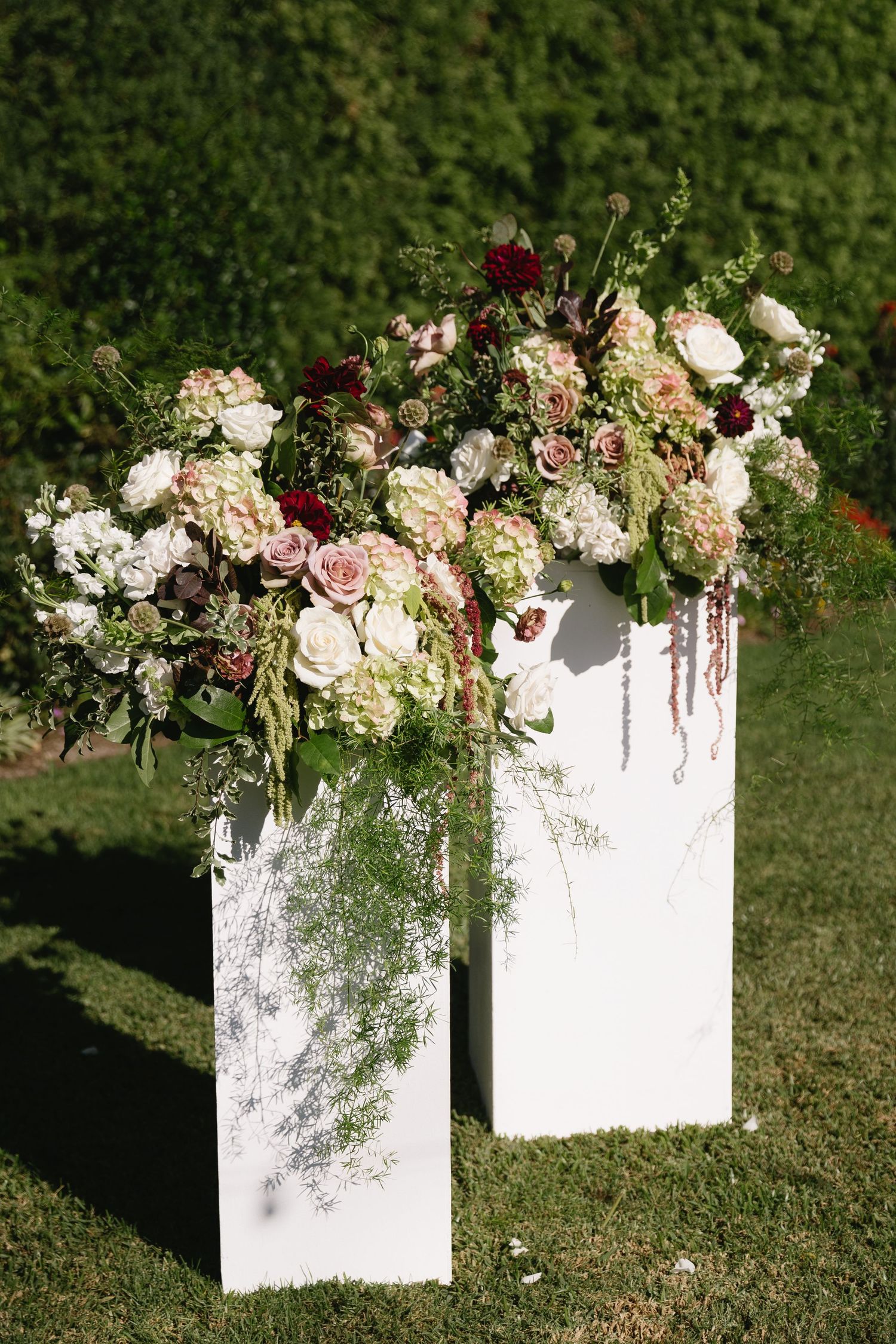 White floral arrangements with blush and burgundy blooms displayed on pedestals against green foliage.