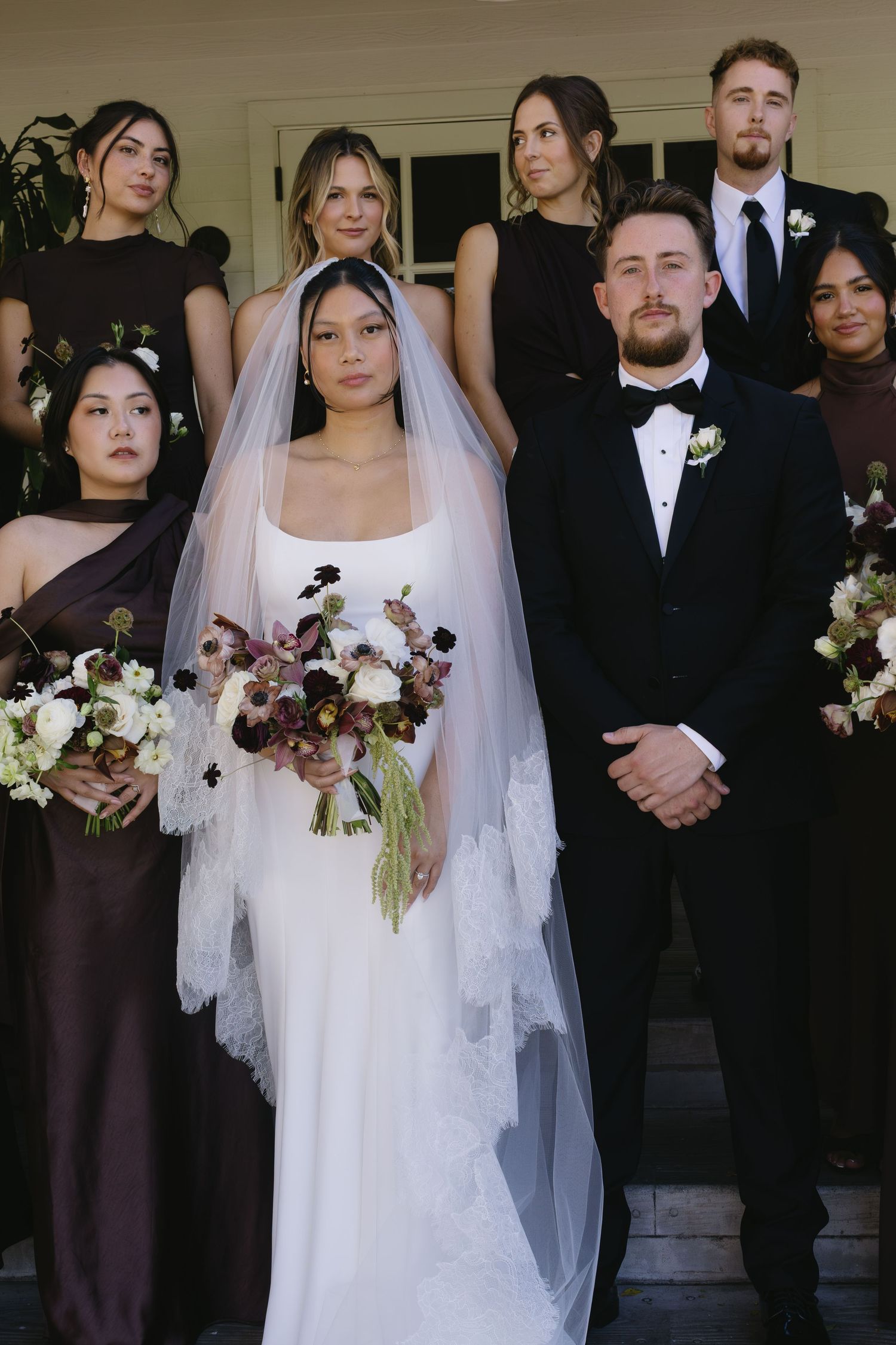 A wedding party poses formally in burgundy dresses and black tuxedos with coordinated floral bouquets.
