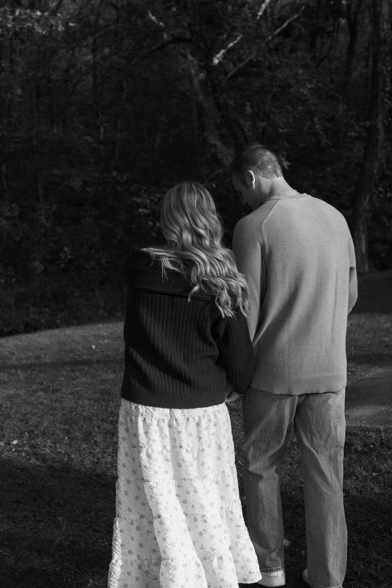 A couple in casual attire stands close together in a black and white outdoor portrait.