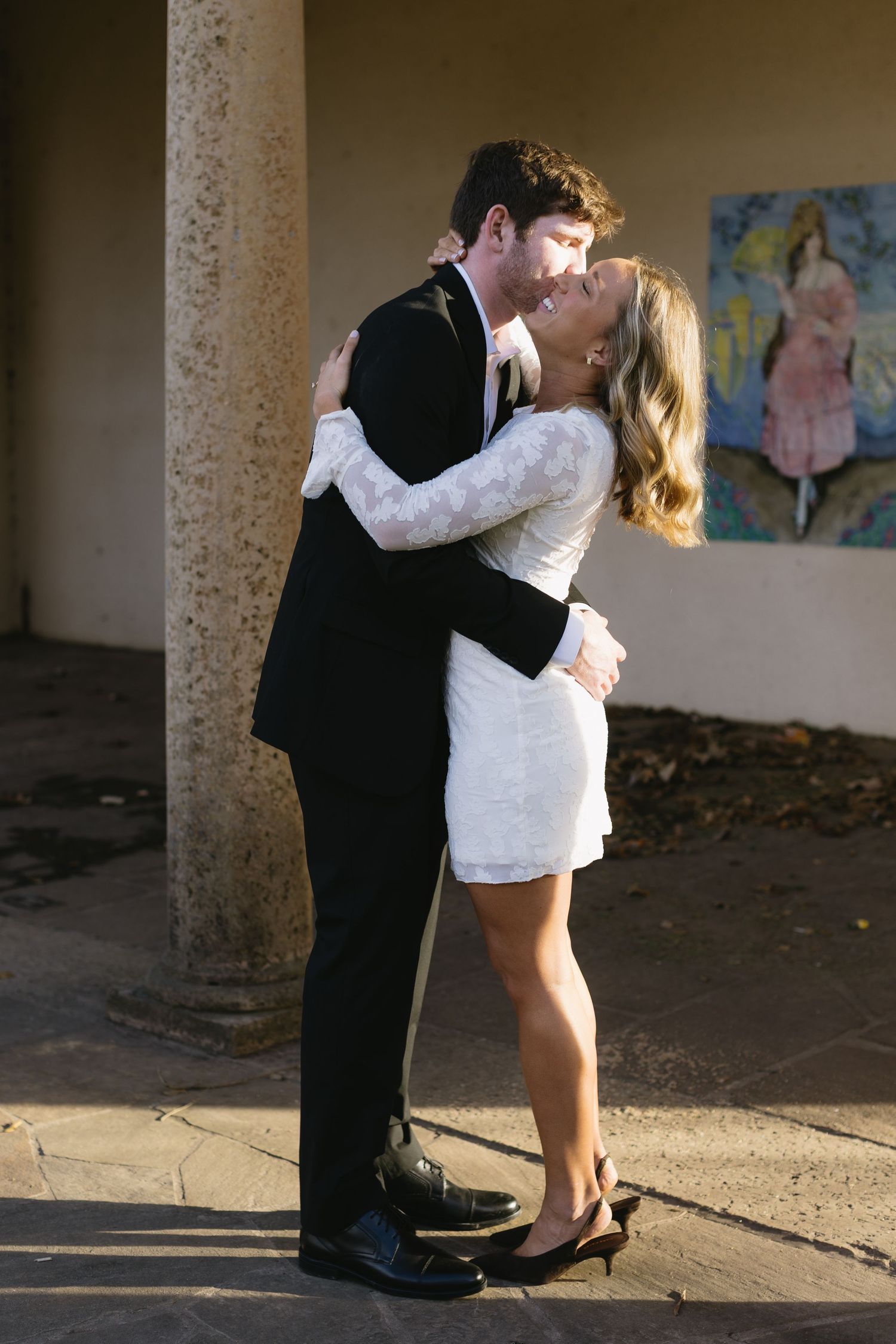 A romantic couple embraces in formal attire against a stone column with artwork in the background.