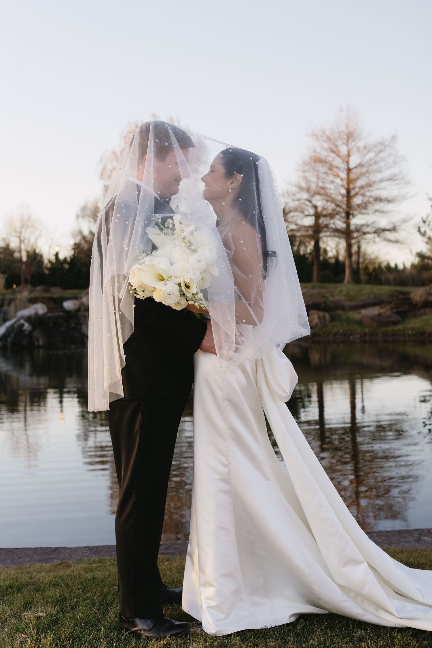 Romantic wedding couple portraits by a lake at sunset with flowing veil and white bouquet.