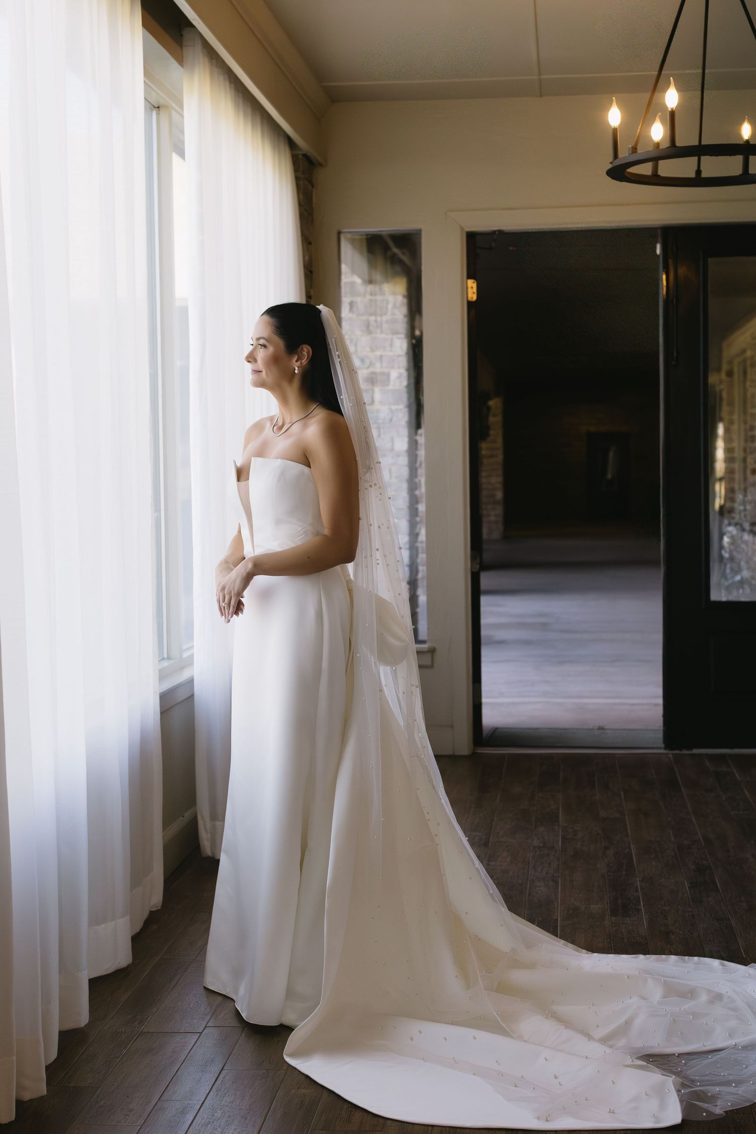 Bride in classic white gown standing by large windows in an elegant indoor setting.
