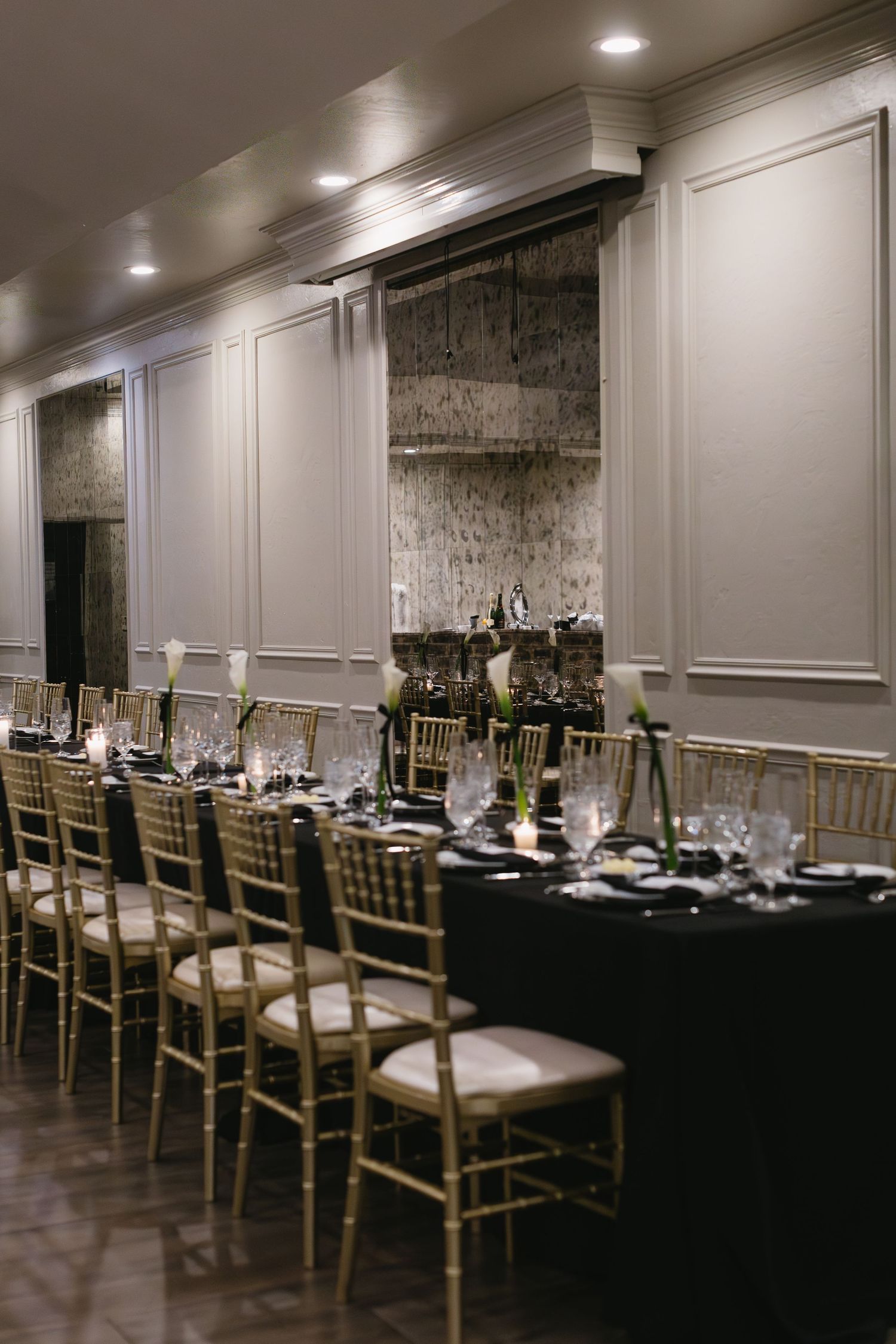 Formal dining setup with gold chairs and black tablecloths in an upscale venue with white paneled walls.