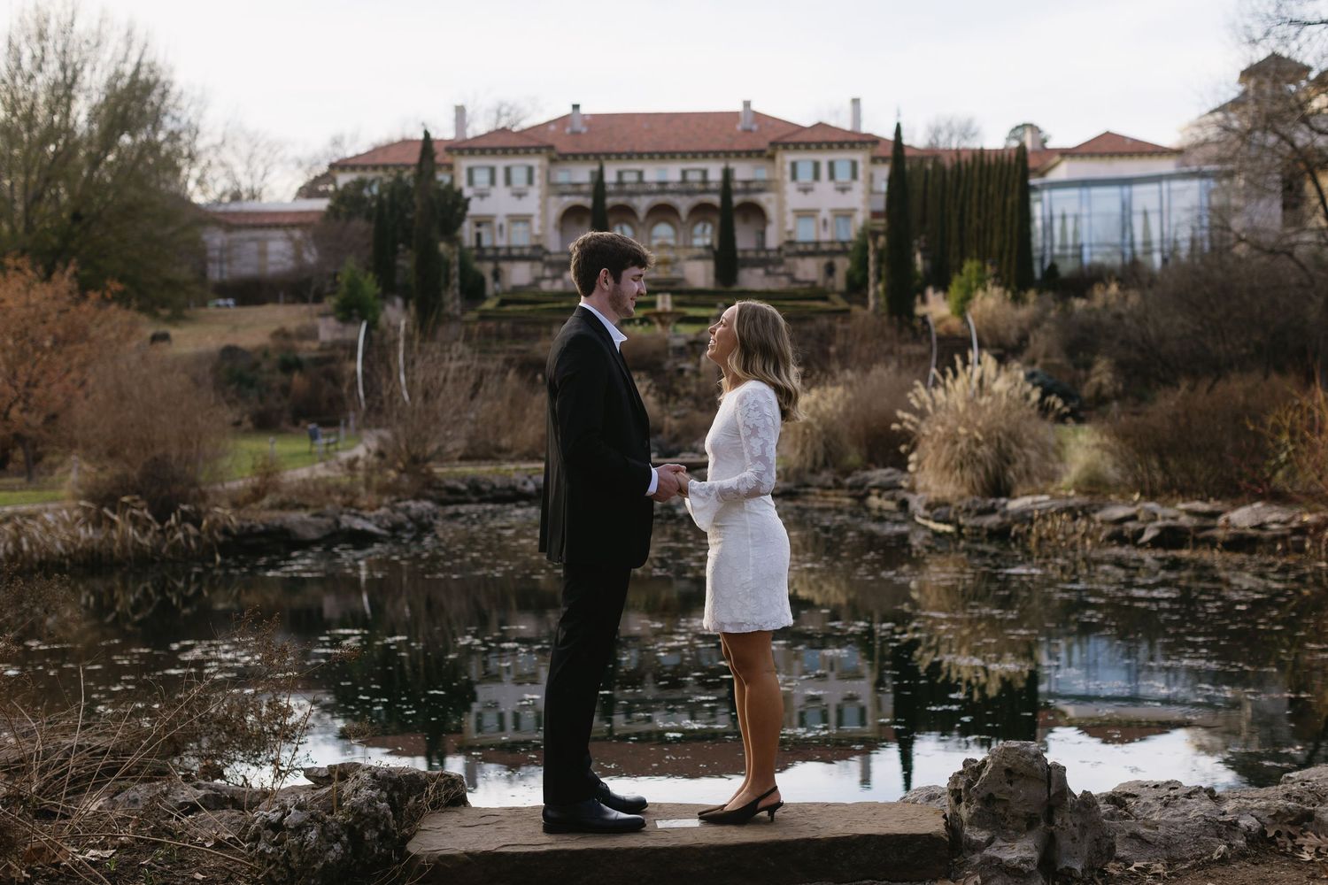 A couple stands together on a wooden dock overlooking a pond with buildings in the background.