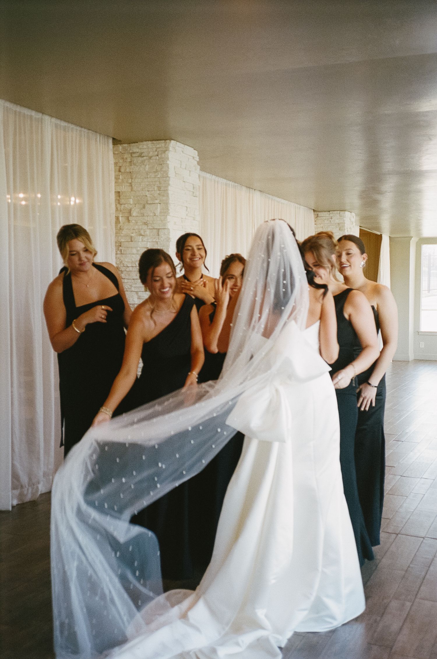 Wedding party in black dresses admiring a flowing white gown in an indoor setting.