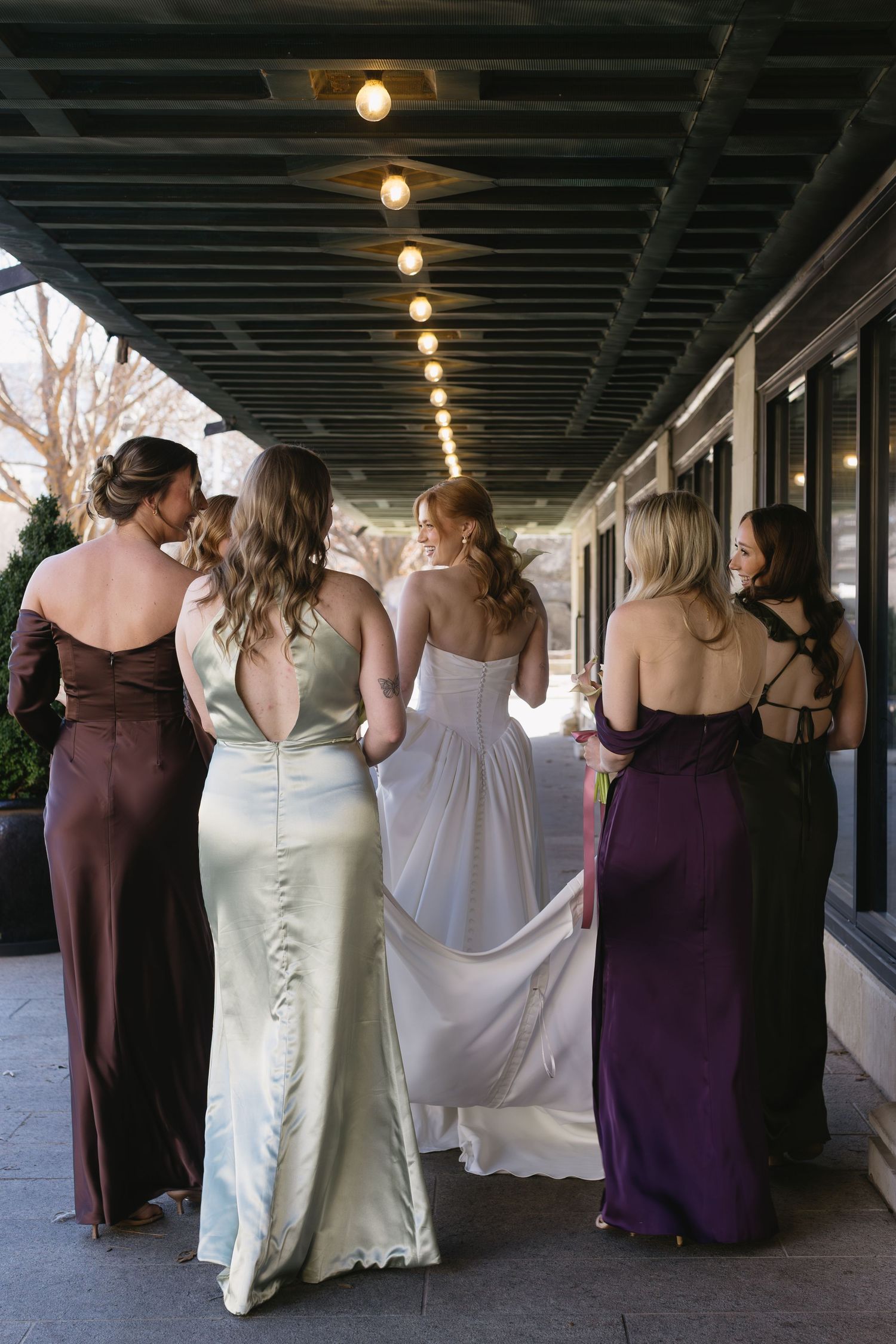 Bridesmaids in burgundy and champagne dresses walk together down a covered walkway.