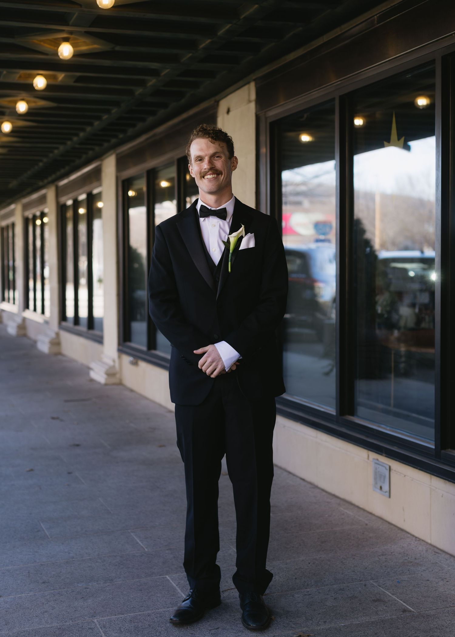A groom in classic black tuxedo stands in front of a commercial storefront.