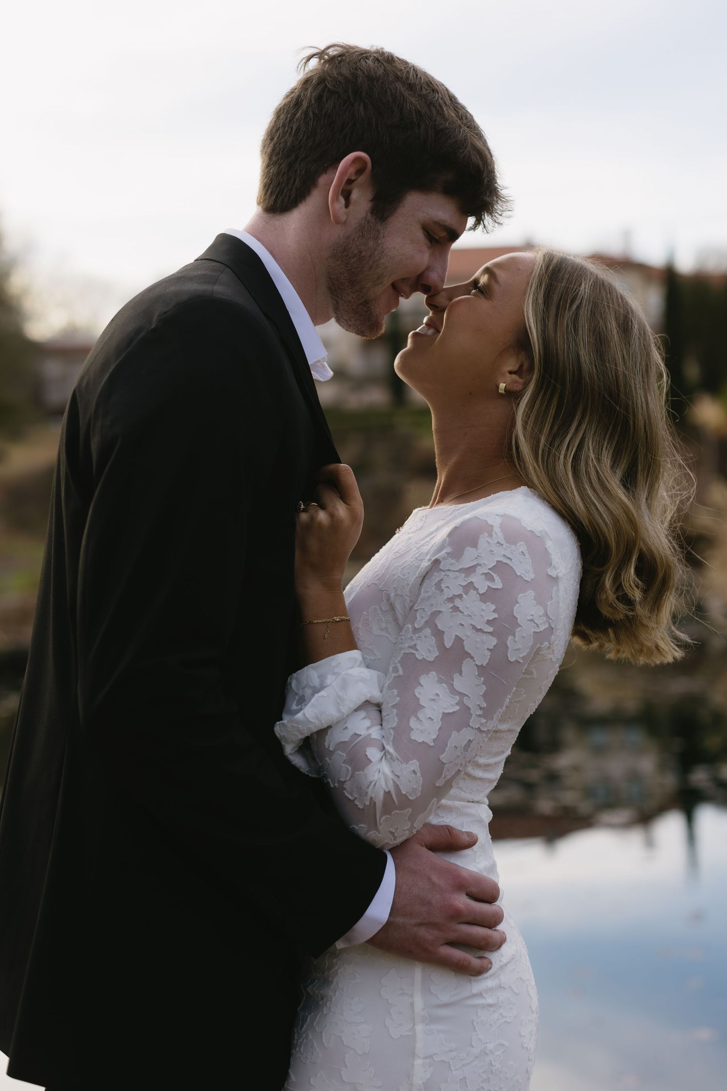 A romantic wedding photo of a couple embracing by a lake, with the bride wearing a lace-sleeved white dress.