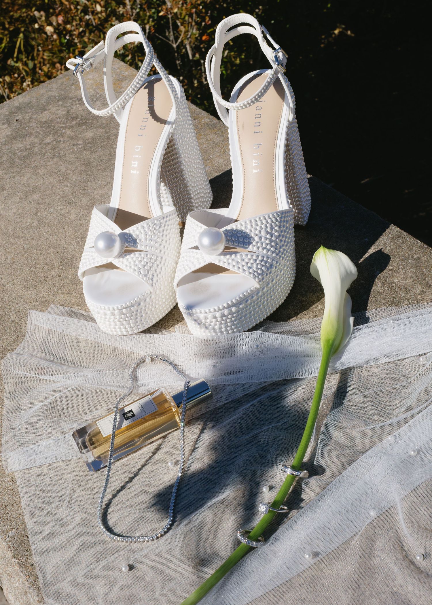 Elegant white bridal shoes with crystals displayed next to a calla lily and perfume bottle on tulle fabric.