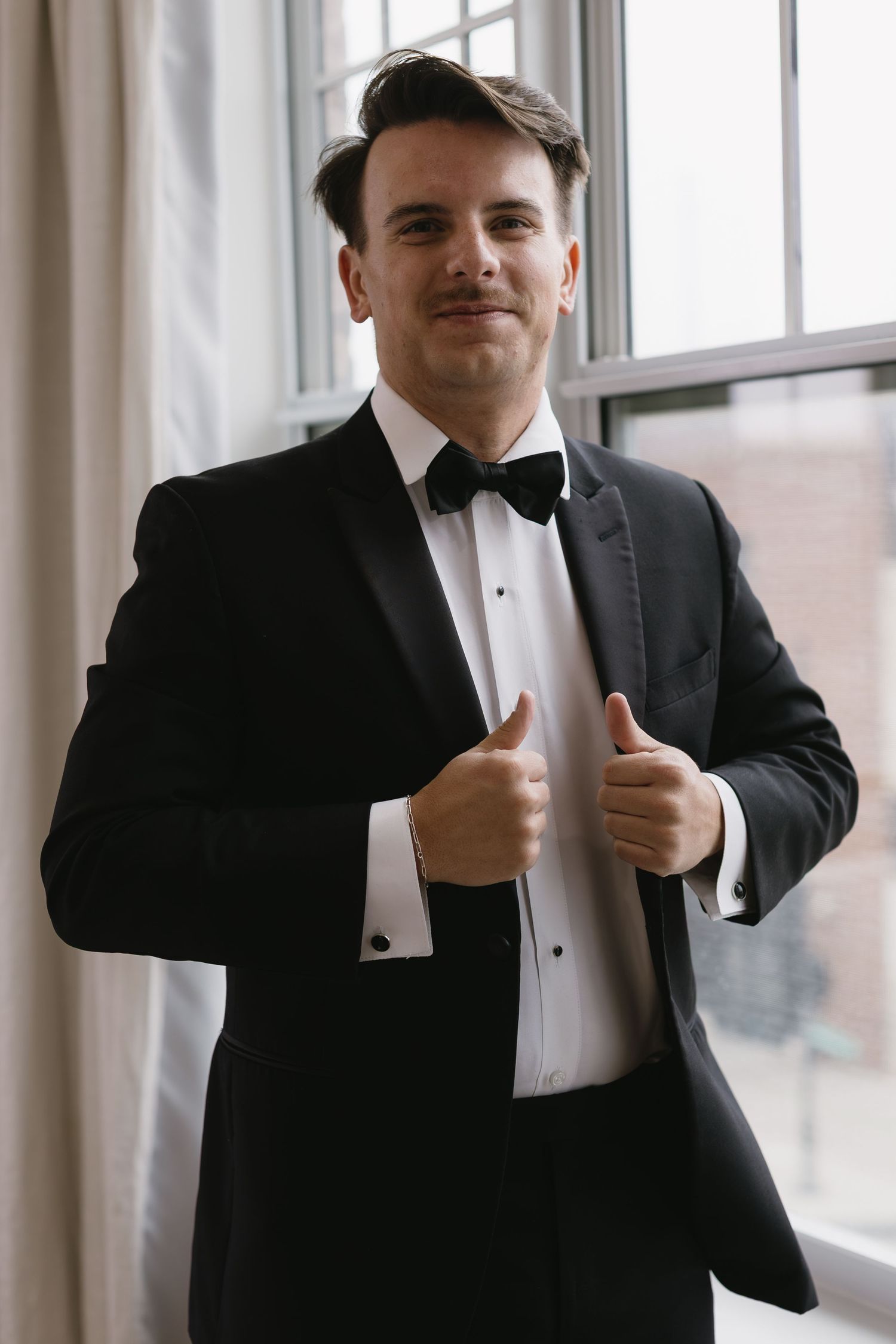A person in a black tuxedo and bow tie gives a thumbs up gesture while standing near bright windows.