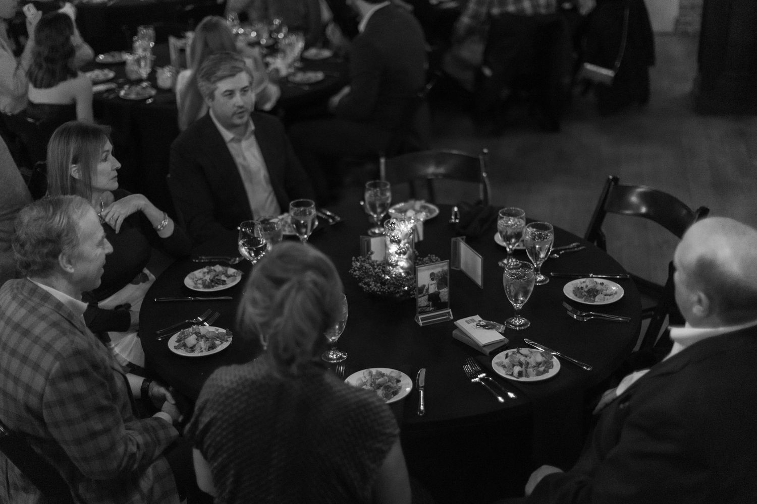 Guests seated at multiple dining tables in a dimly lit restaurant with plates of food and drinks.