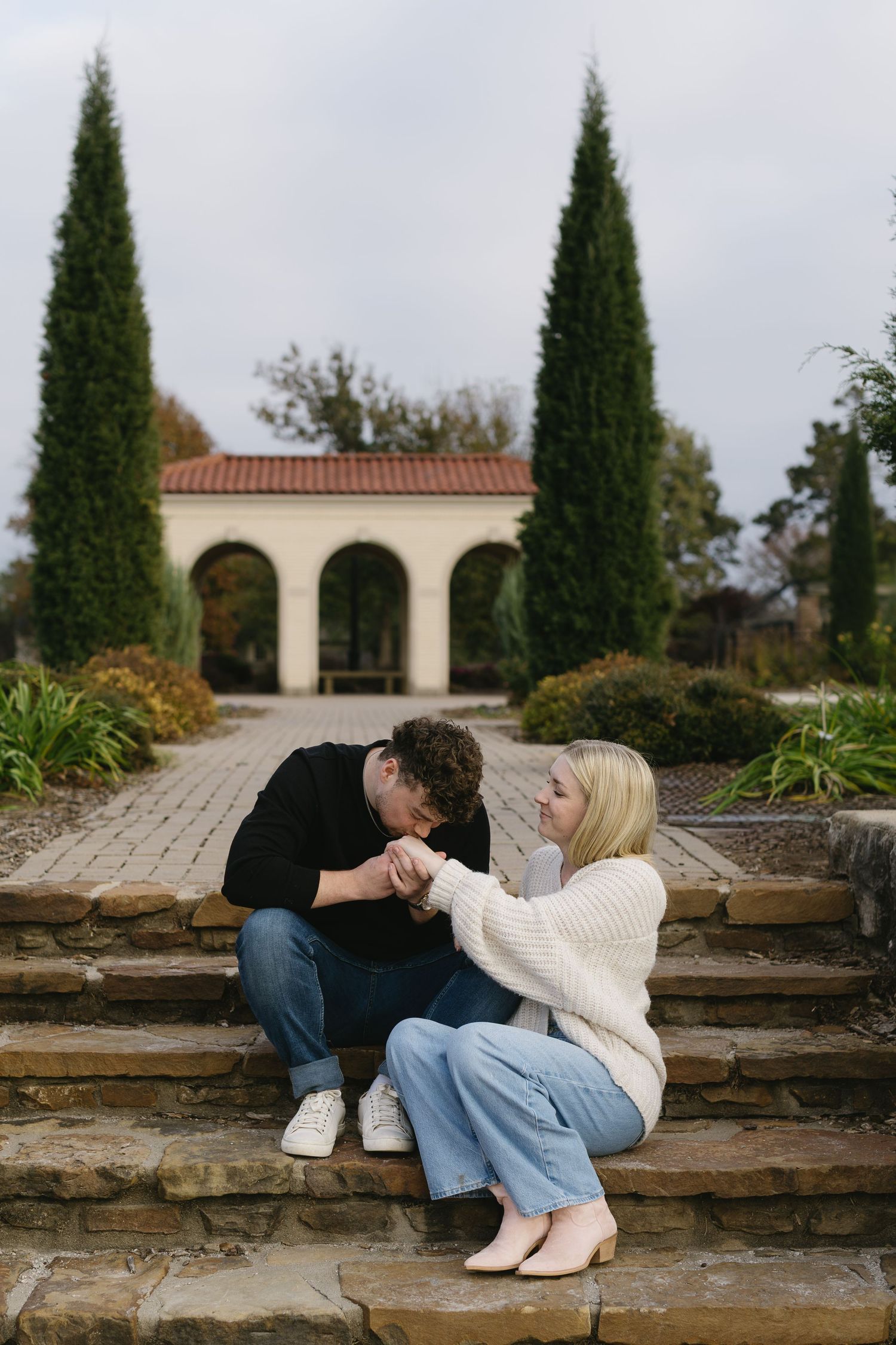 A couple sits on stone steps surrounded by cypress trees and arched architecture in a romantic garden setting.