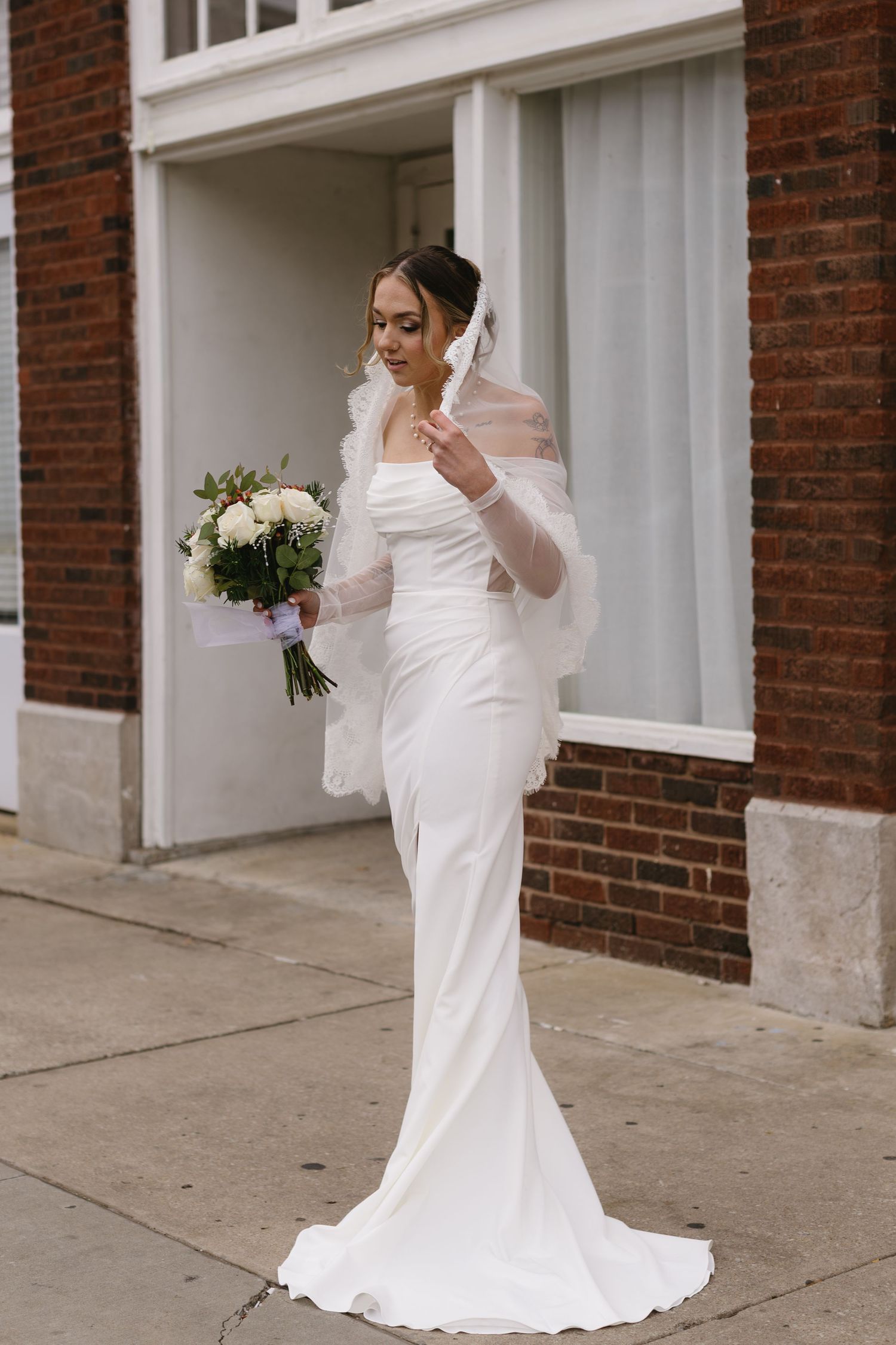 Elegant bride in fitted white wedding gown poses on sidewalk outside brick building.