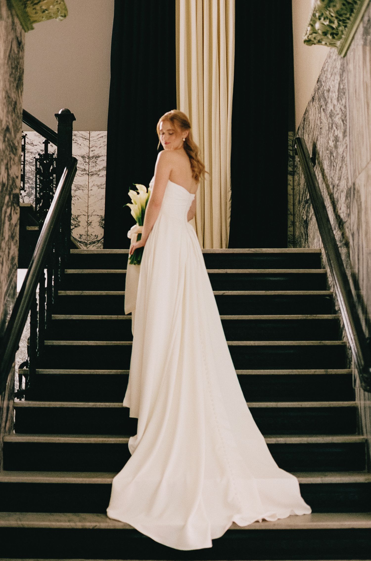 A bride in a flowing white gown ascends marble stairs while holding a bouquet.