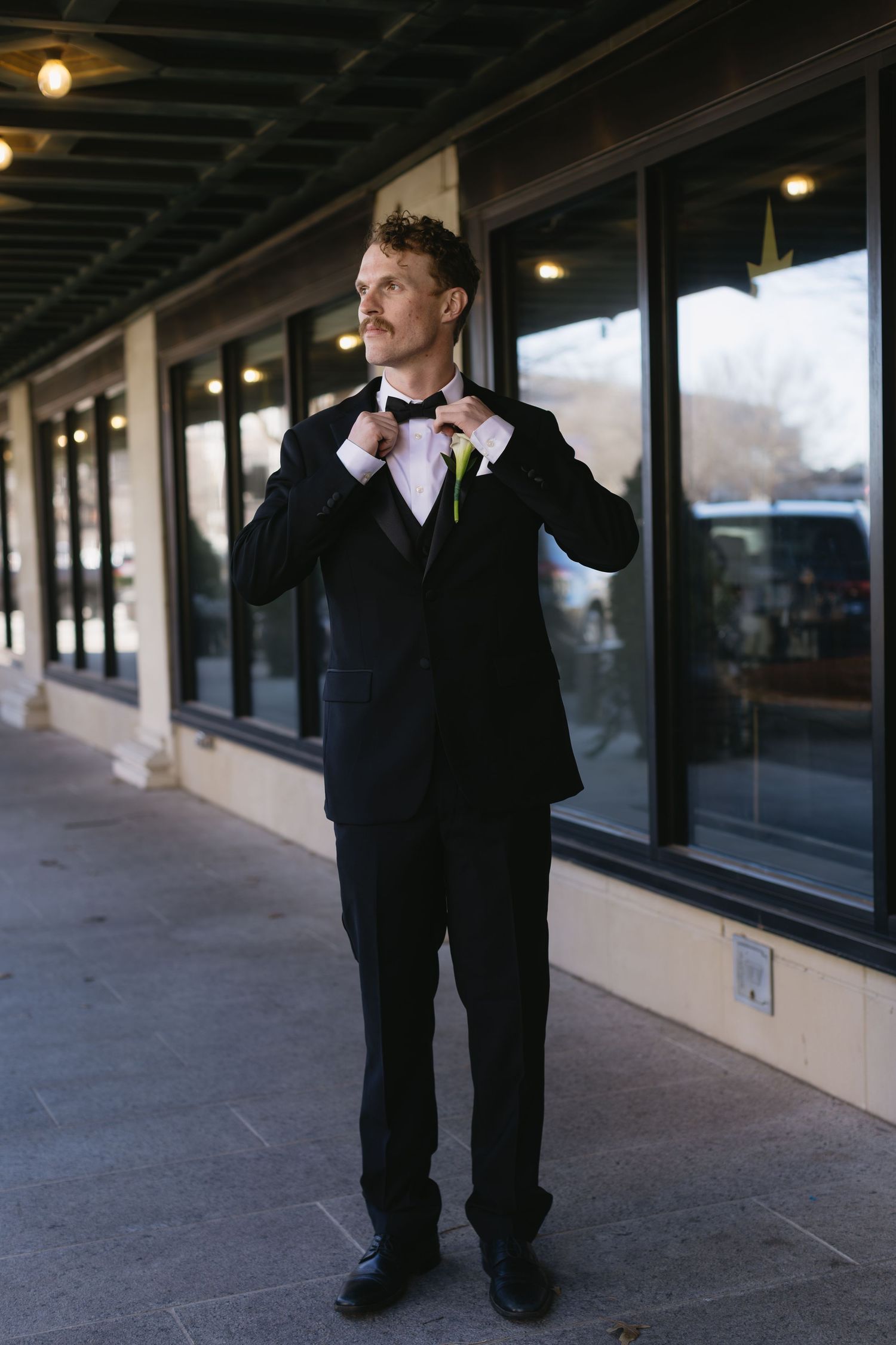 Groom in formal attire stands thoughtfully along a covered walkway with windows.